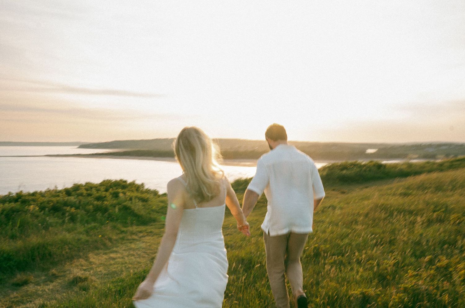 A couple walks through a sunlit meadow at golden hour with rolling hills in Halifax, Nova Scotia.