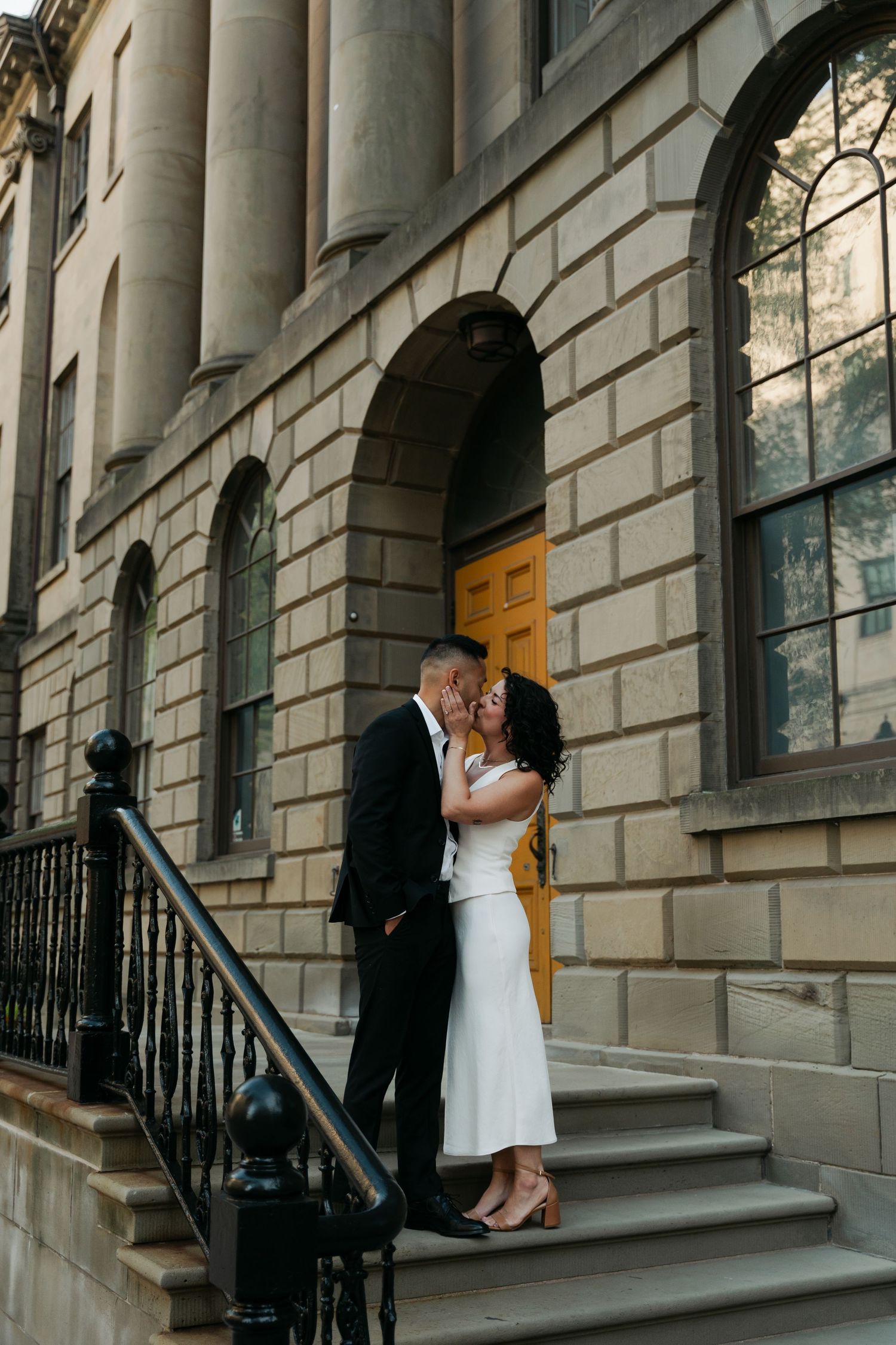 A couple shares an intimate moment on stone steps outside a classic building with yellow doors in downtown Halifax, Nova Scotia.