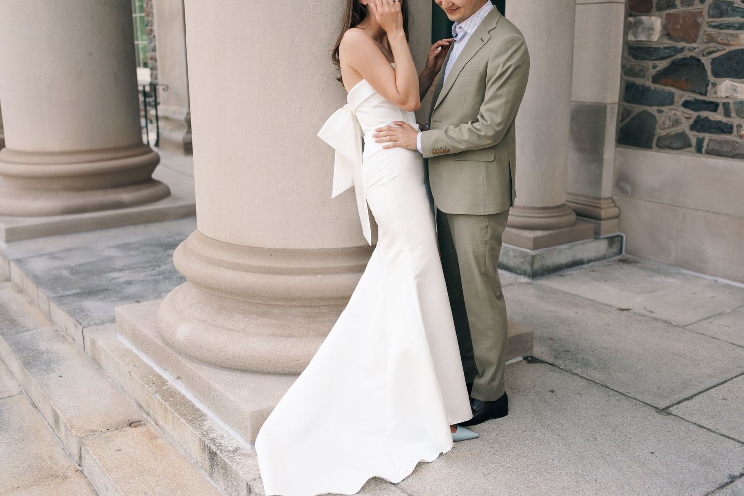 A couple embraces in formal wedding attire next to stone columns, with the bride wearing a sleek white gown in Halifax, Nova Scotia.