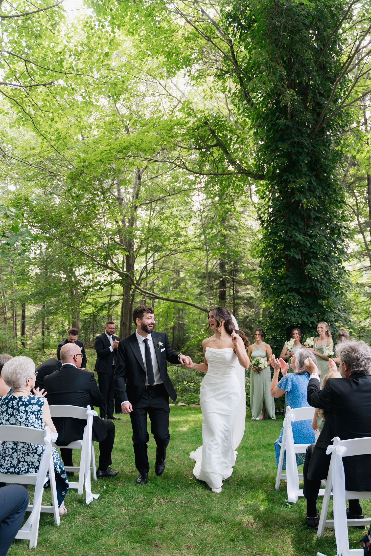 Wedding guests celebrate at an outdoor garden ceremony with white chairs and blue accents among lush green trees in Halifax, Nova Scotia.