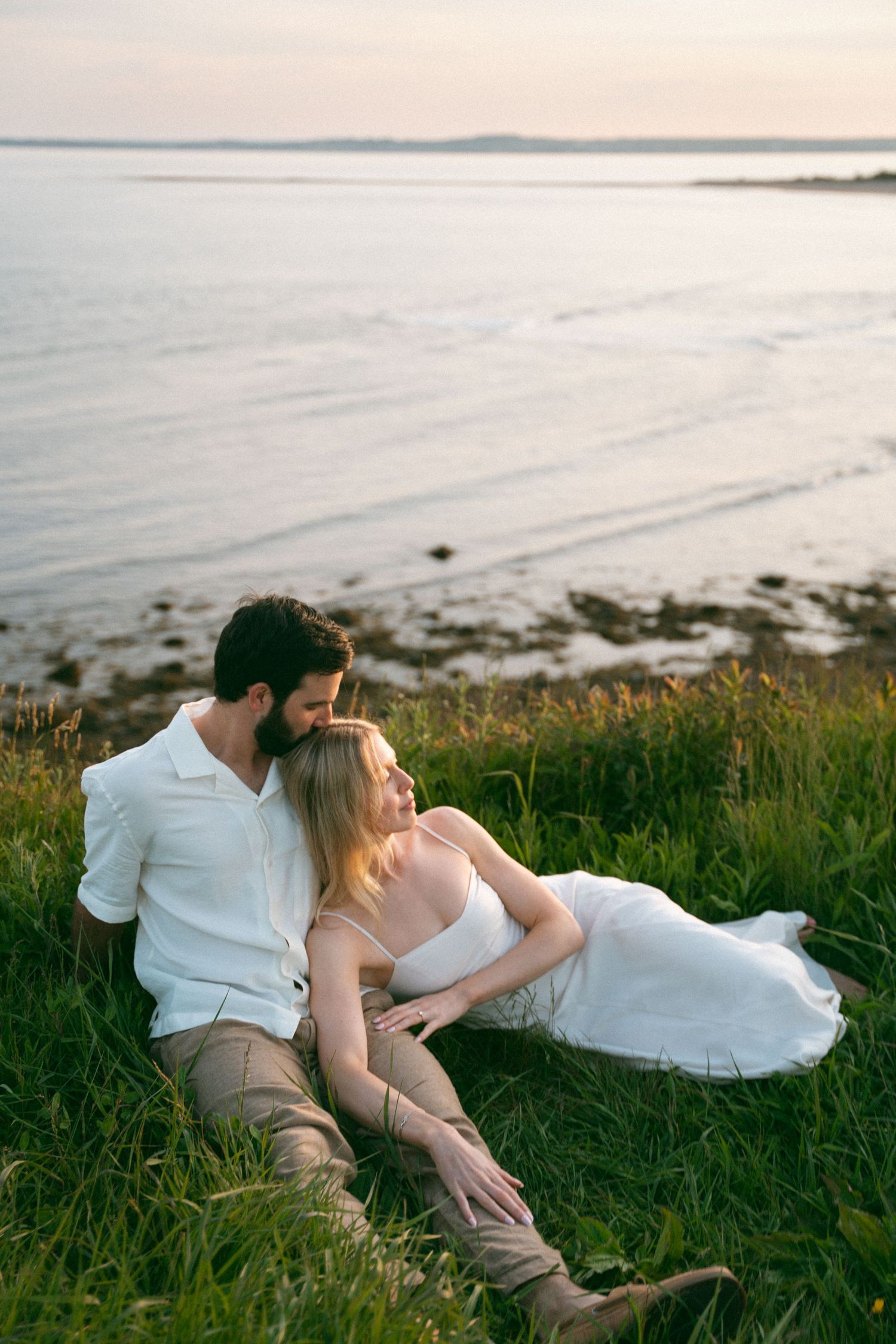 A couple in white clothing shares an intimate moment on a grassy hillside overlooking a serene ocean at sunset in Halifax, Nova Scotia.