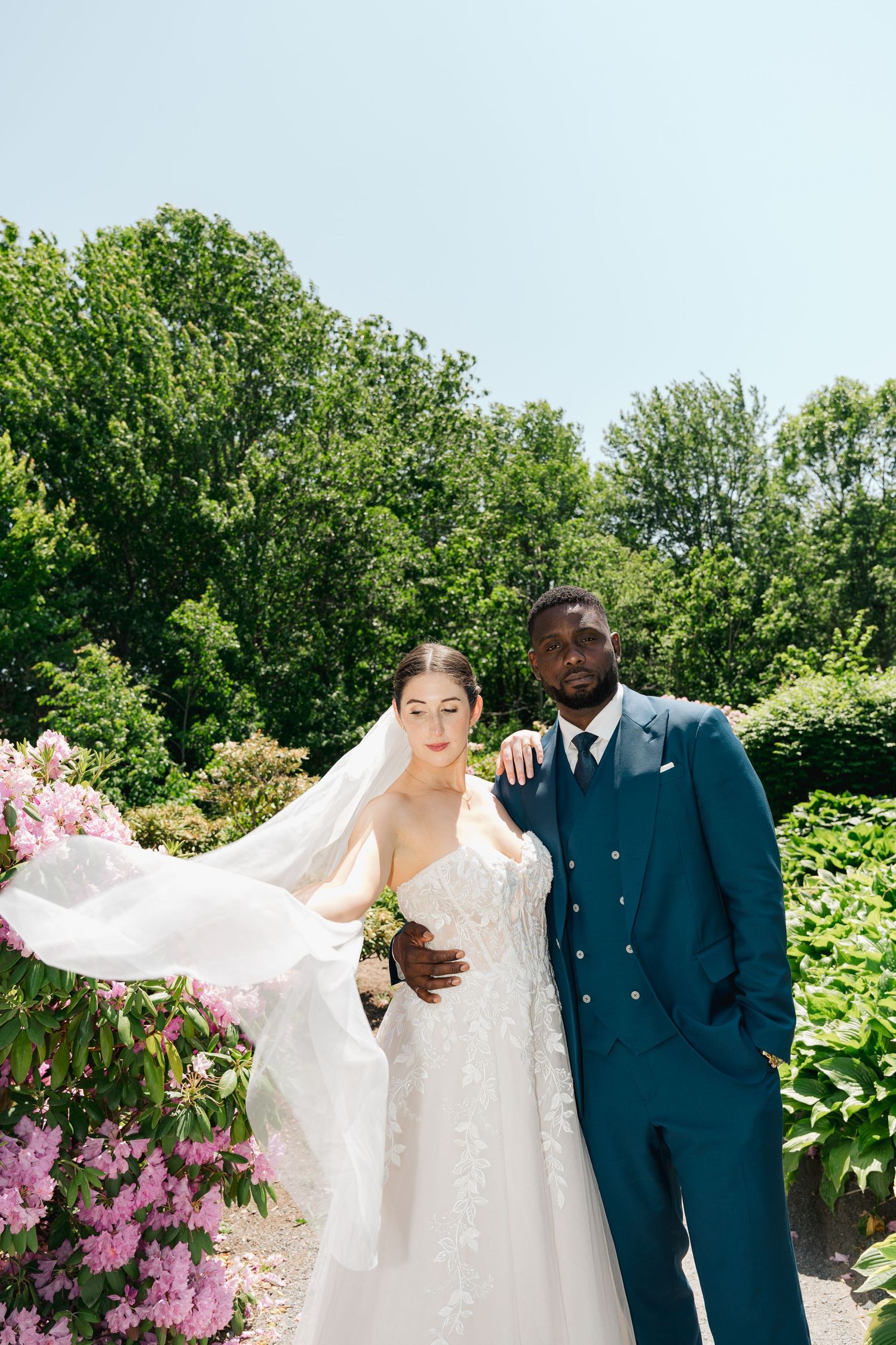 A wedding couple poses outdoors with flowing veil against lush green garden and purple flowers in summer in Halifax, Nova Scotia.