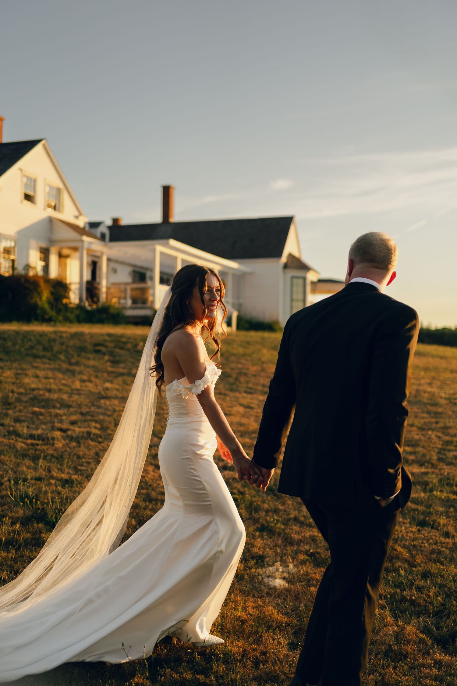 A bride's flowing veil catches the golden hour light as she walks across a grassy field in Halifax, Nova Scotia.