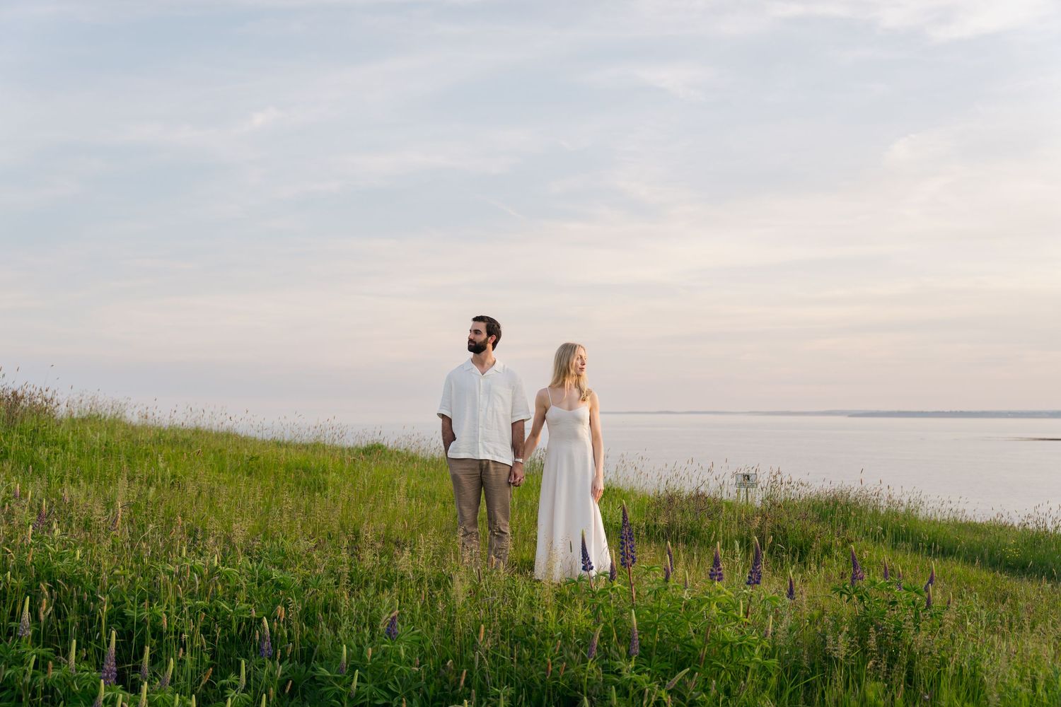 A couple walks together through a lush green field in Halifax, Nova Scotia with a serene waterfront view at sunset.