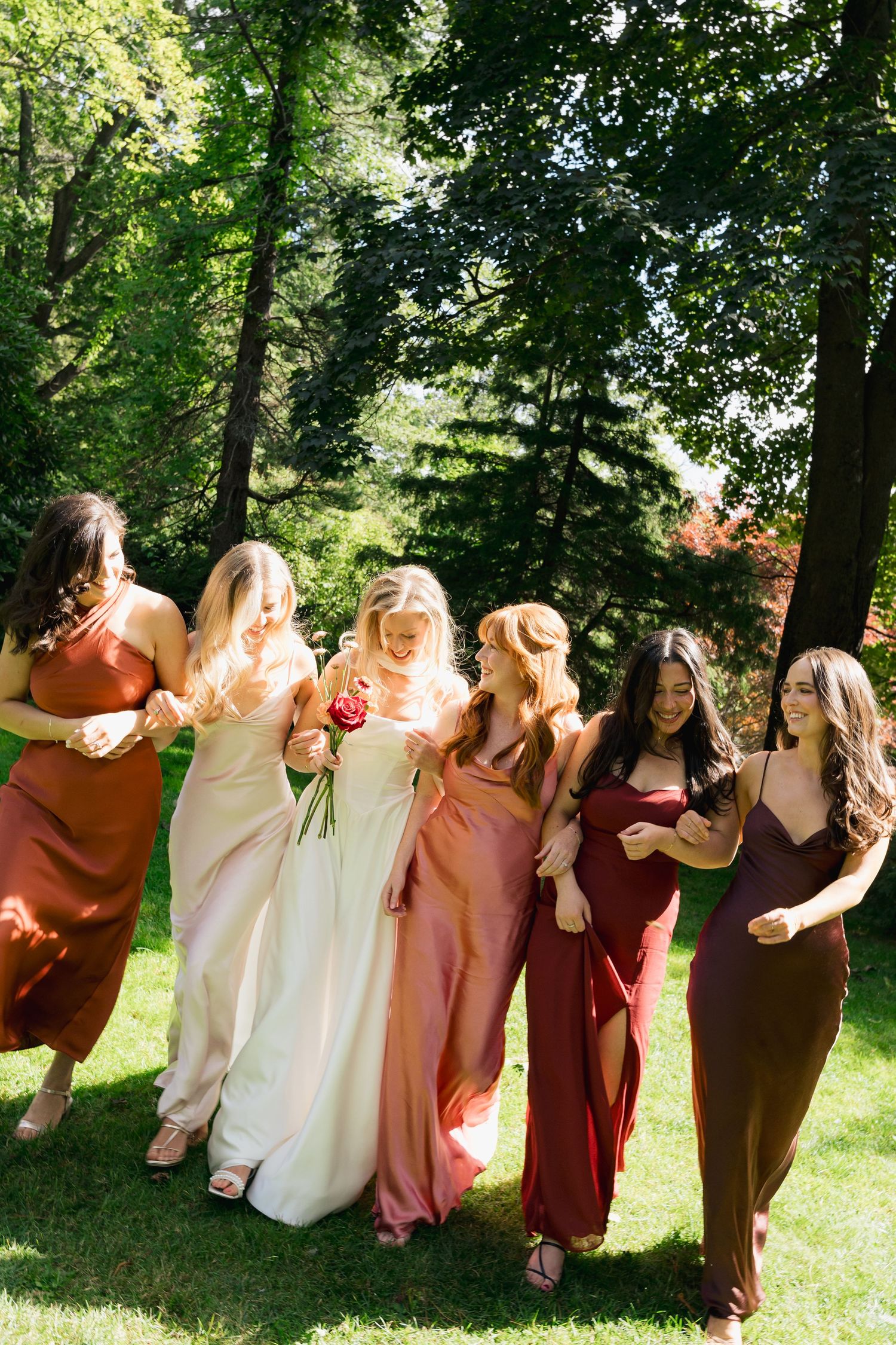 Bridesmaids in earth-toned dresses pose together outdoors in a sunny garden setting in Halifax, Nova Scotia.