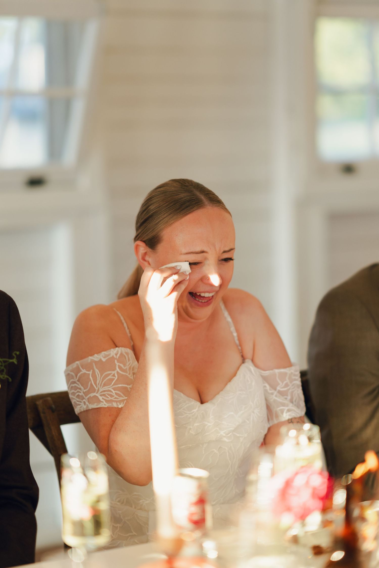 A person in a white lace dress wipes away tears while laughing during an emotional wedding reception moment in Nova Scotia.