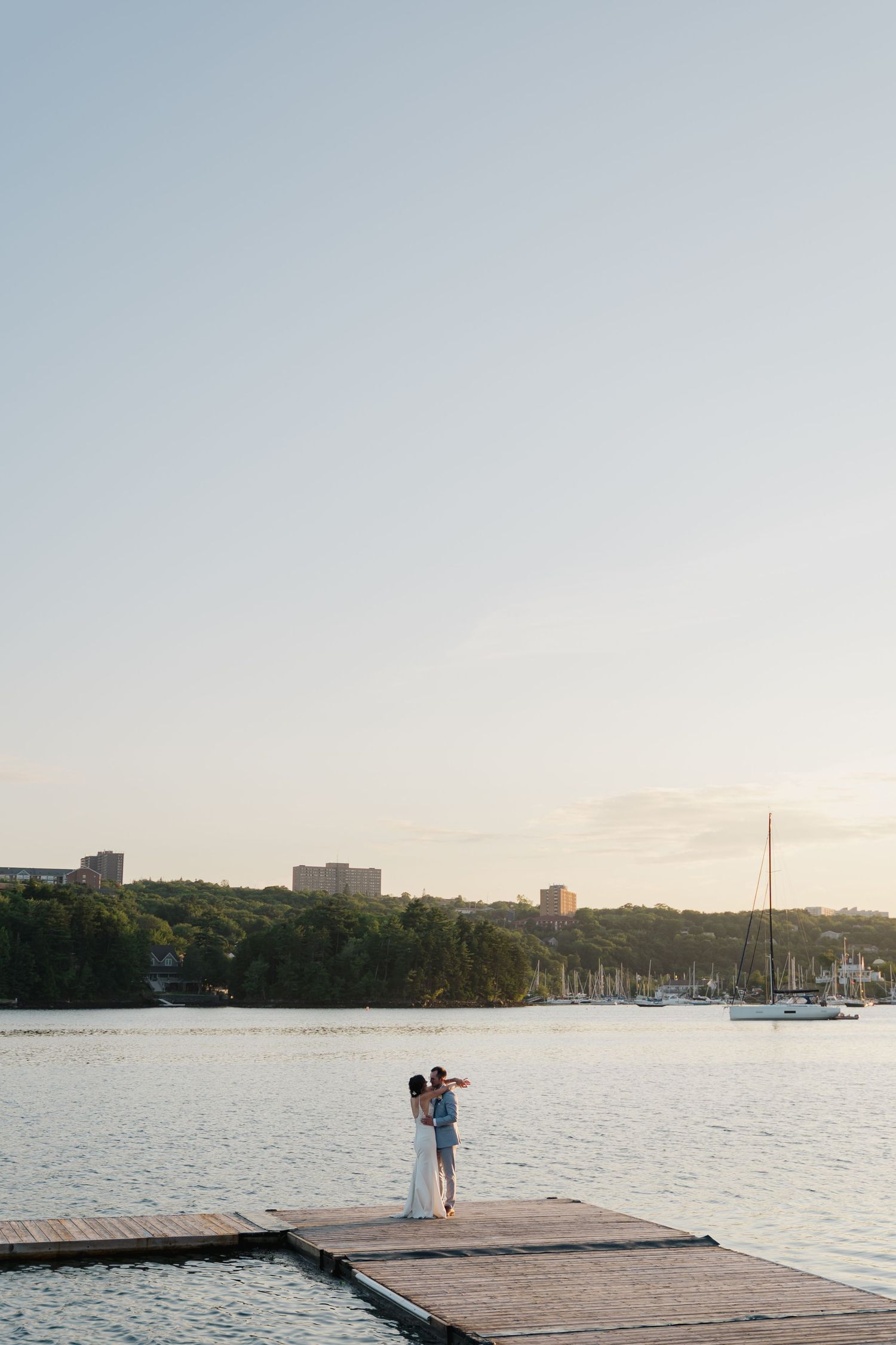 Wedding couple poses on a wooden dock at sunset overlooking a serene harbor with boats and city skyline in Halifax, Nova Scotia.