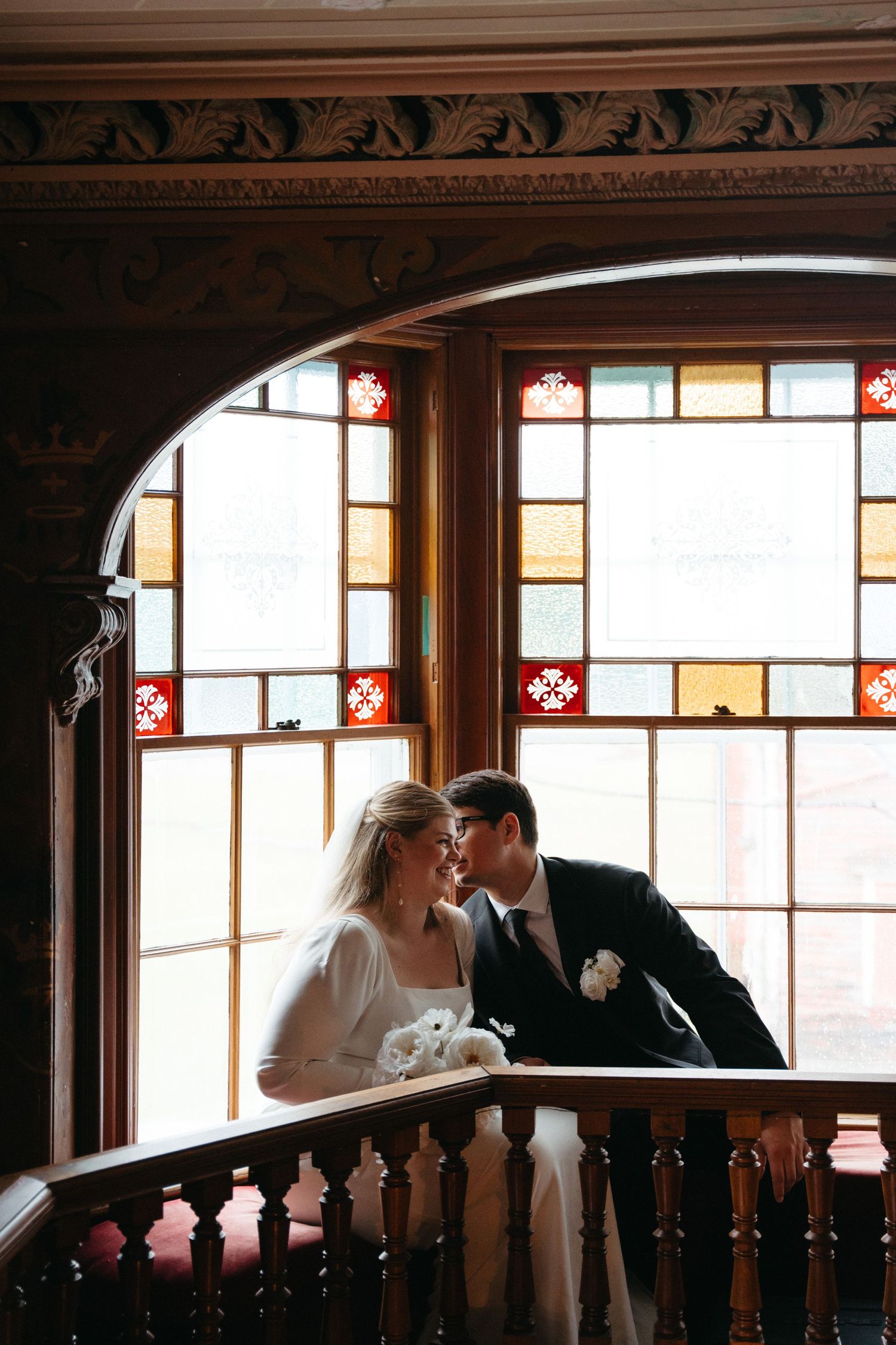 A romantic wedding scene shows a couple sharing intimate moments along an ornate wooden railing in warm indoor lighting at Churchill House.