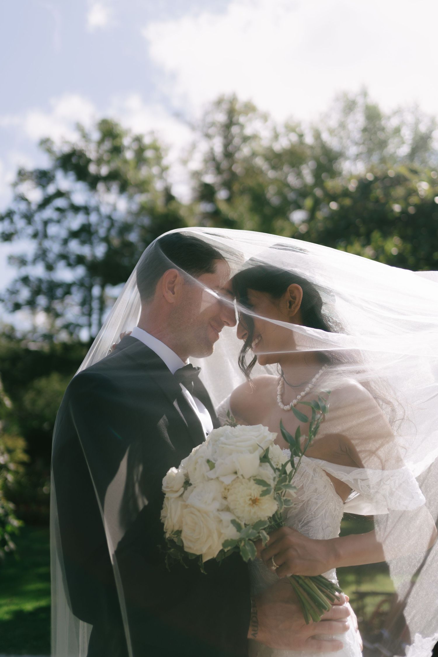 Bride and groom share a romantic moment under a flowing veil with white roses at Halifax Public Garden in Halifax, Nova Scotia.