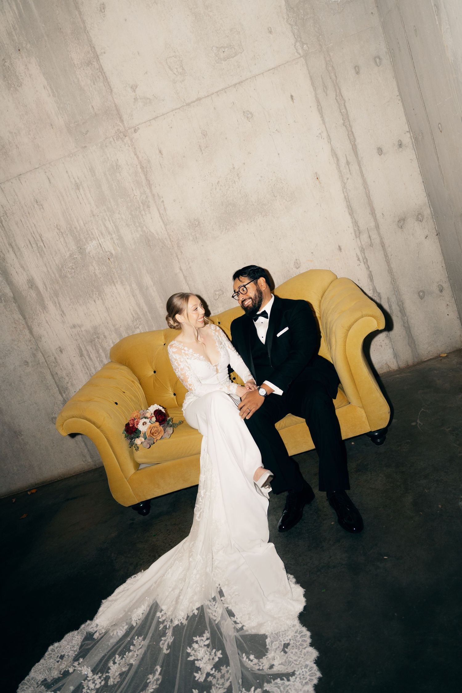 A couple poses together on a yellow vintage sofa against an industrial concrete wall at Lightfoot & Wolfville.