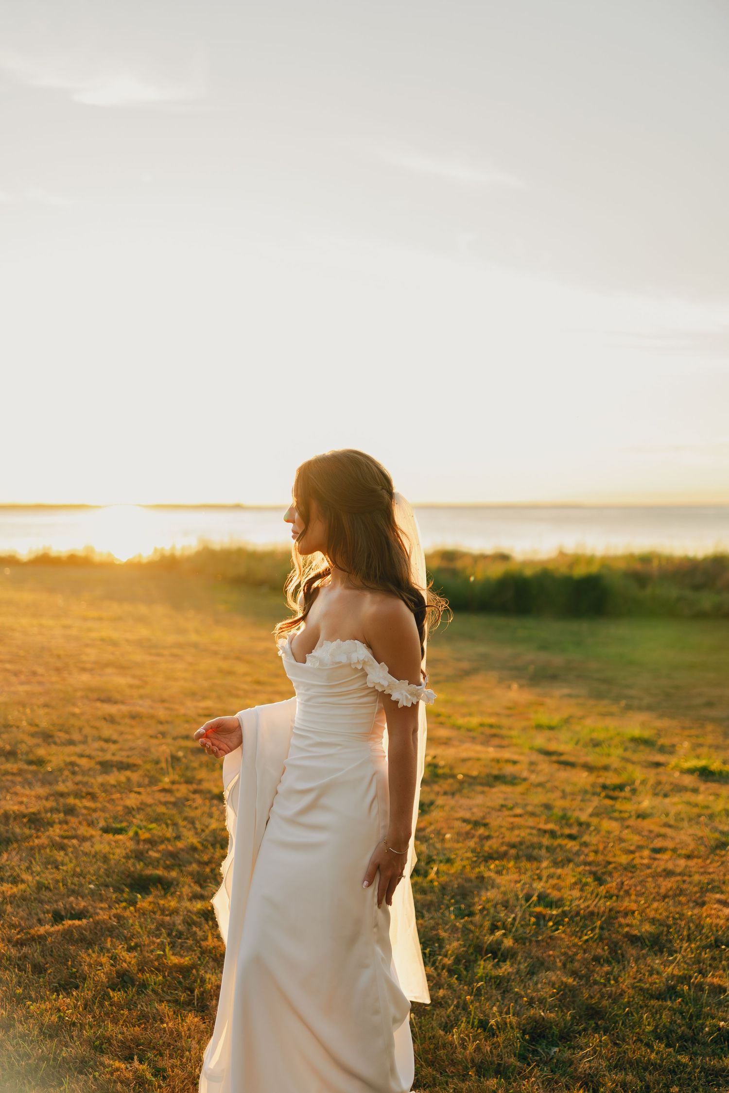 Bride in white dress walking alone across sunlit field during golden hour in Halifax, Nova Scotia.