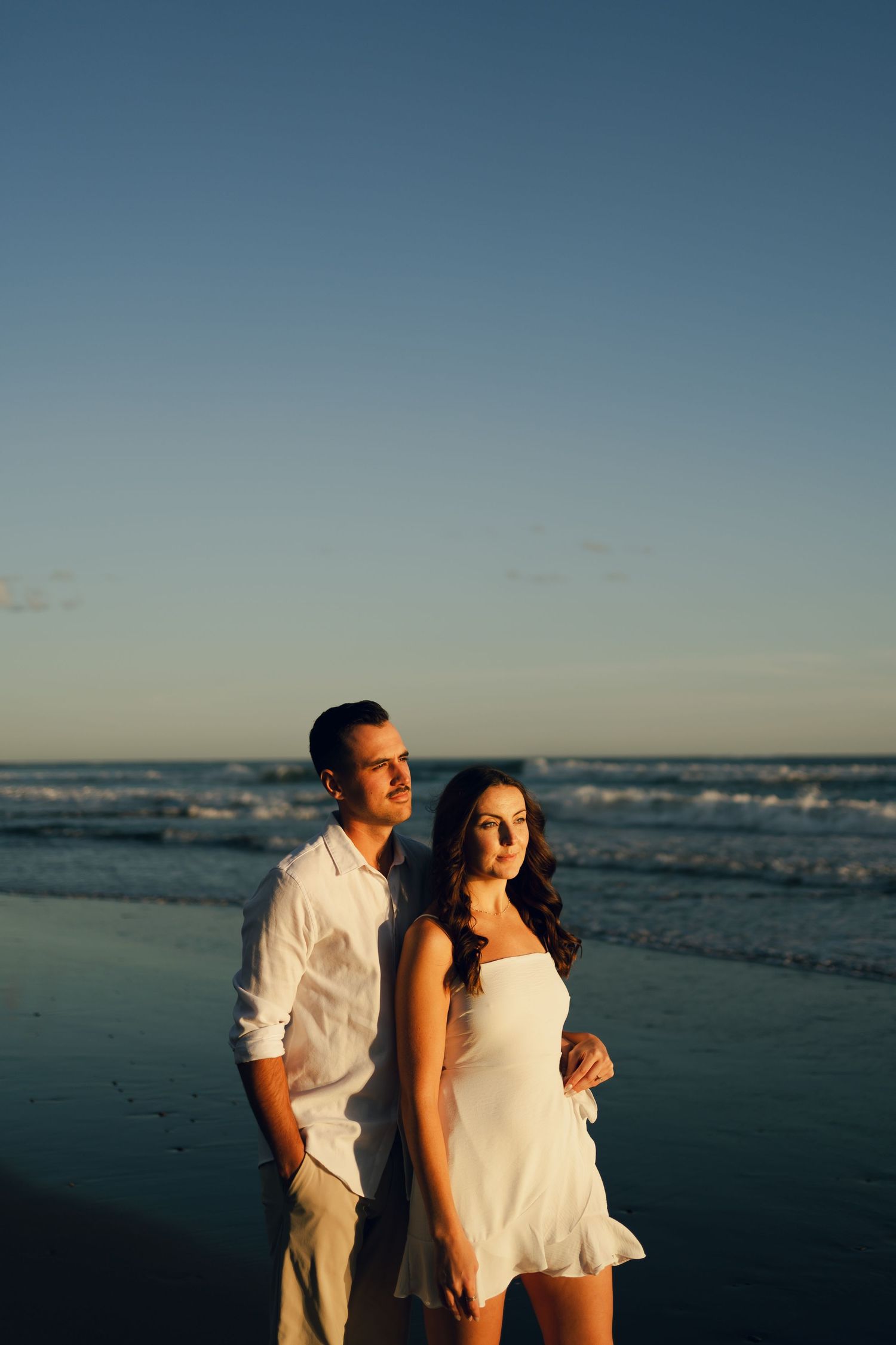 A couple in white outfits shares a romantic moment on a beach in Halifax, Nova Scotia at sunset with ocean waves in the background.