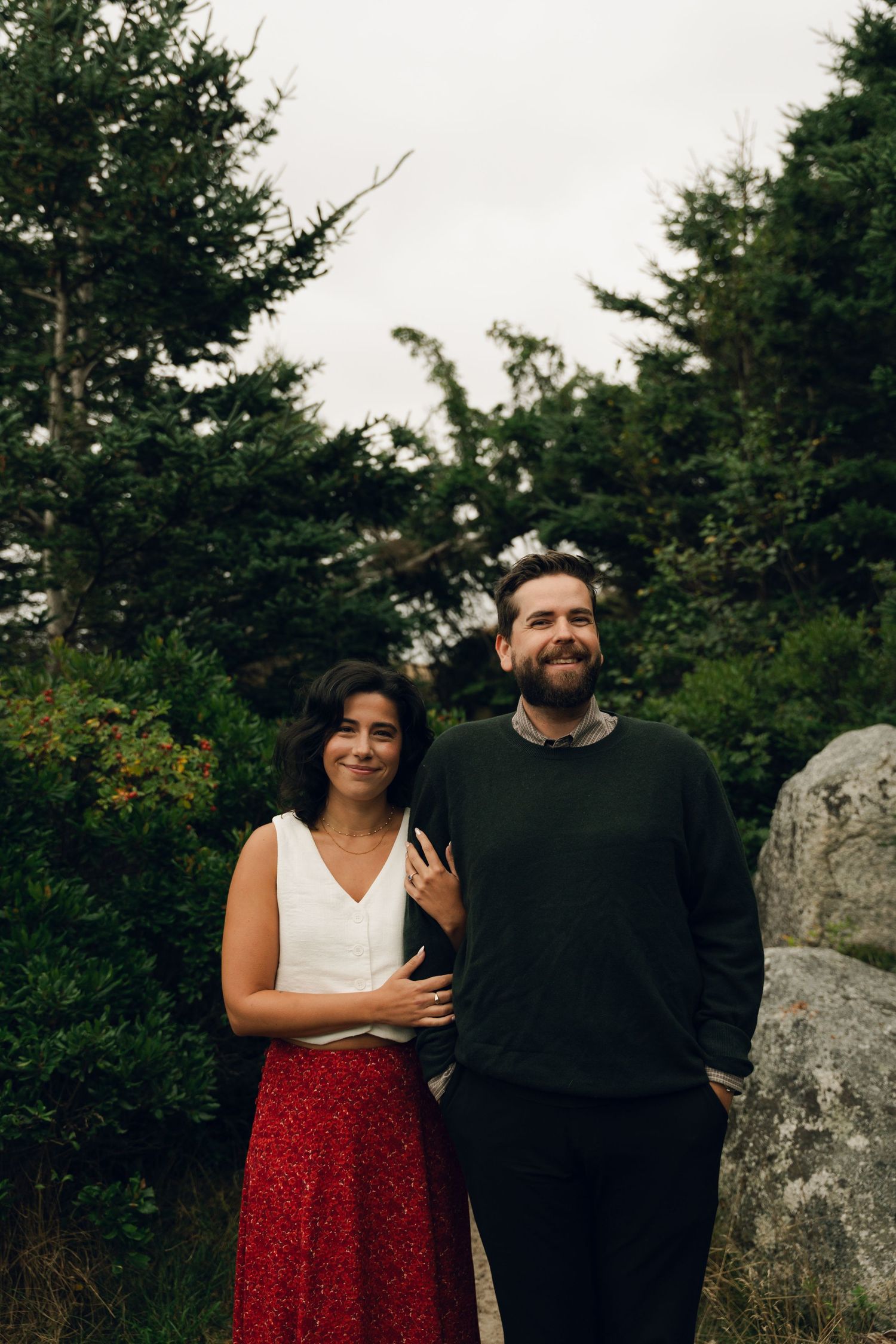 A couple stands together outdoors against evergreen trees and rocks in Halifax, Nova Scotia, with one wearing a red skirt and white top.