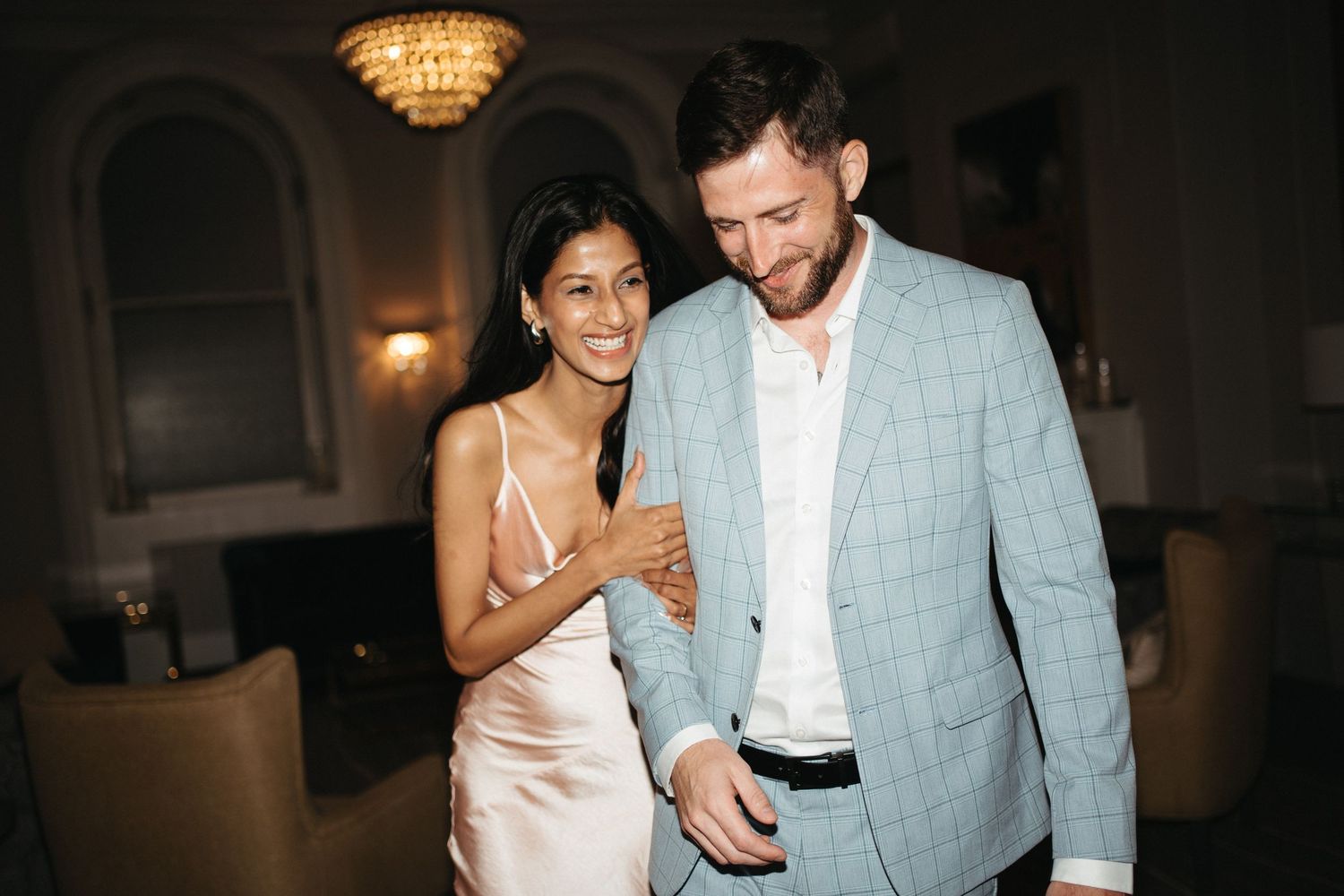 A couple laughs together at an evening event, with the woman wearing a silk dress and man in a light blue blazer in Halifax, Nova Scotia.