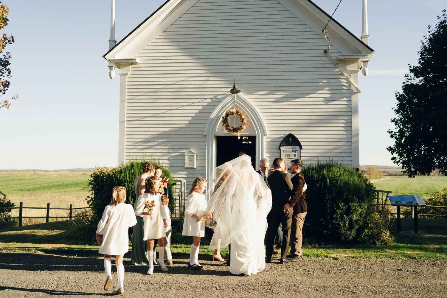 Wedding party poses in sequence outside white country church with steeple on sunny day in Nova Scotia