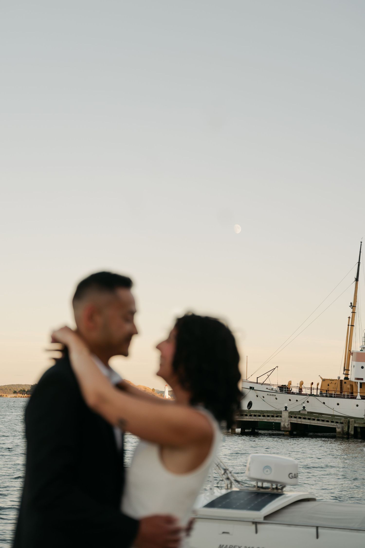 A romantic couple embraces on a boat dock at sunset with a marina and sailboats in the background in waterfront Halifax, Nova Scotia.