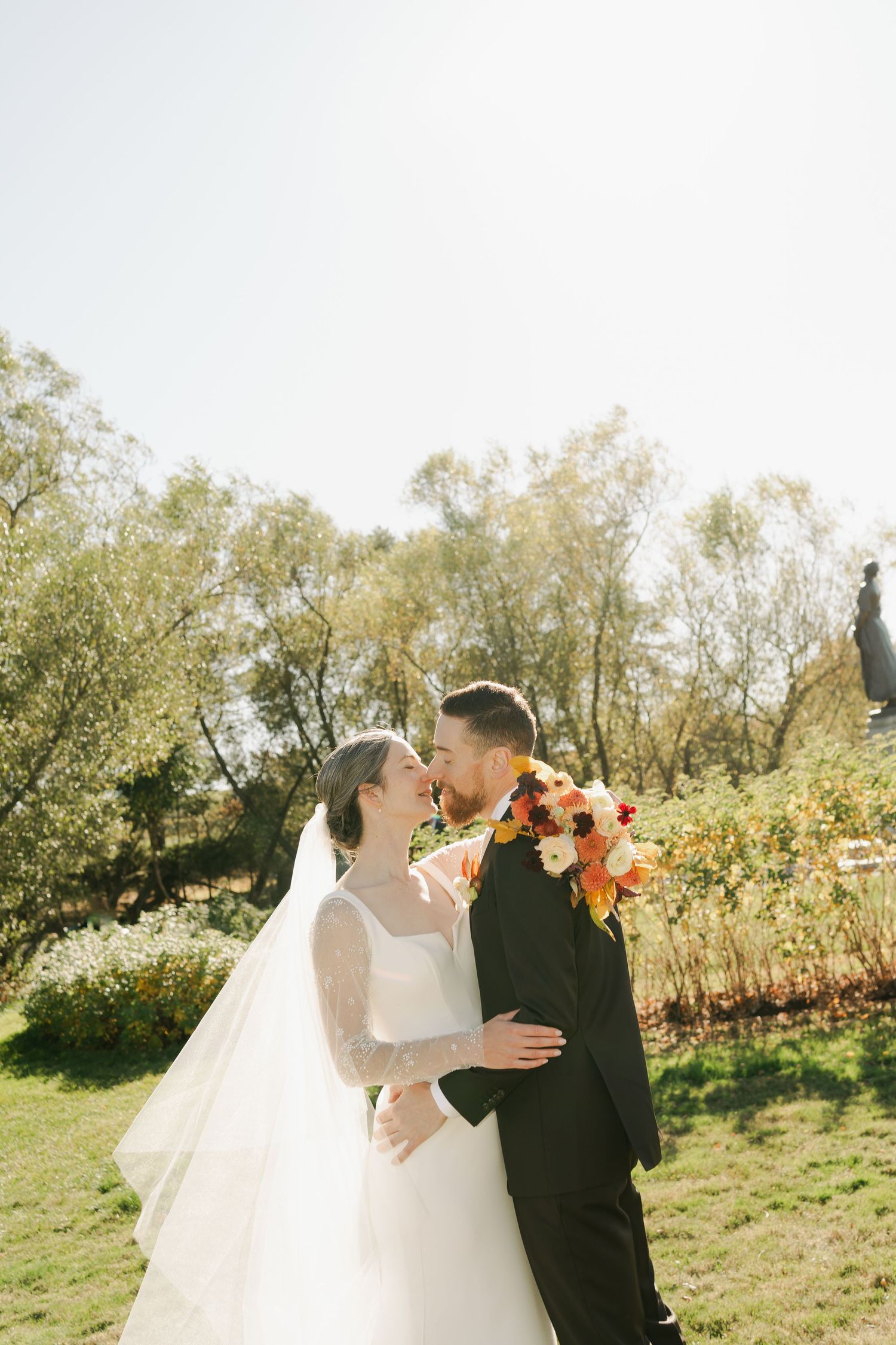 A romantic wedding couple shares intimate moments in a sunlit field with autumn trees in the background in Nova Scotia.