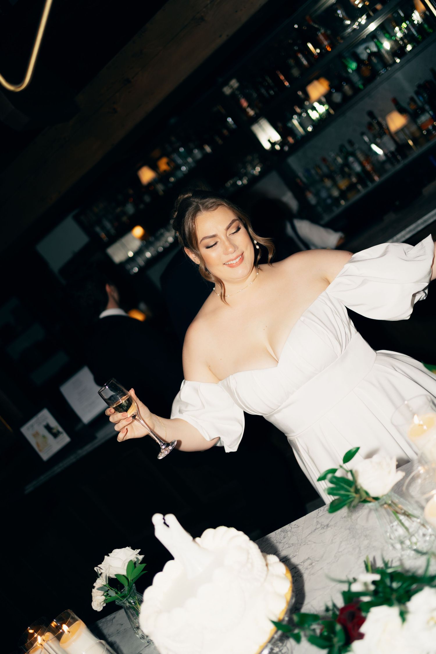 A bride in a white off-shoulder gown cuts into a wedding cake decorated with greenery at Lord Nelson Hotel, Halifax, Nova Scotia.
