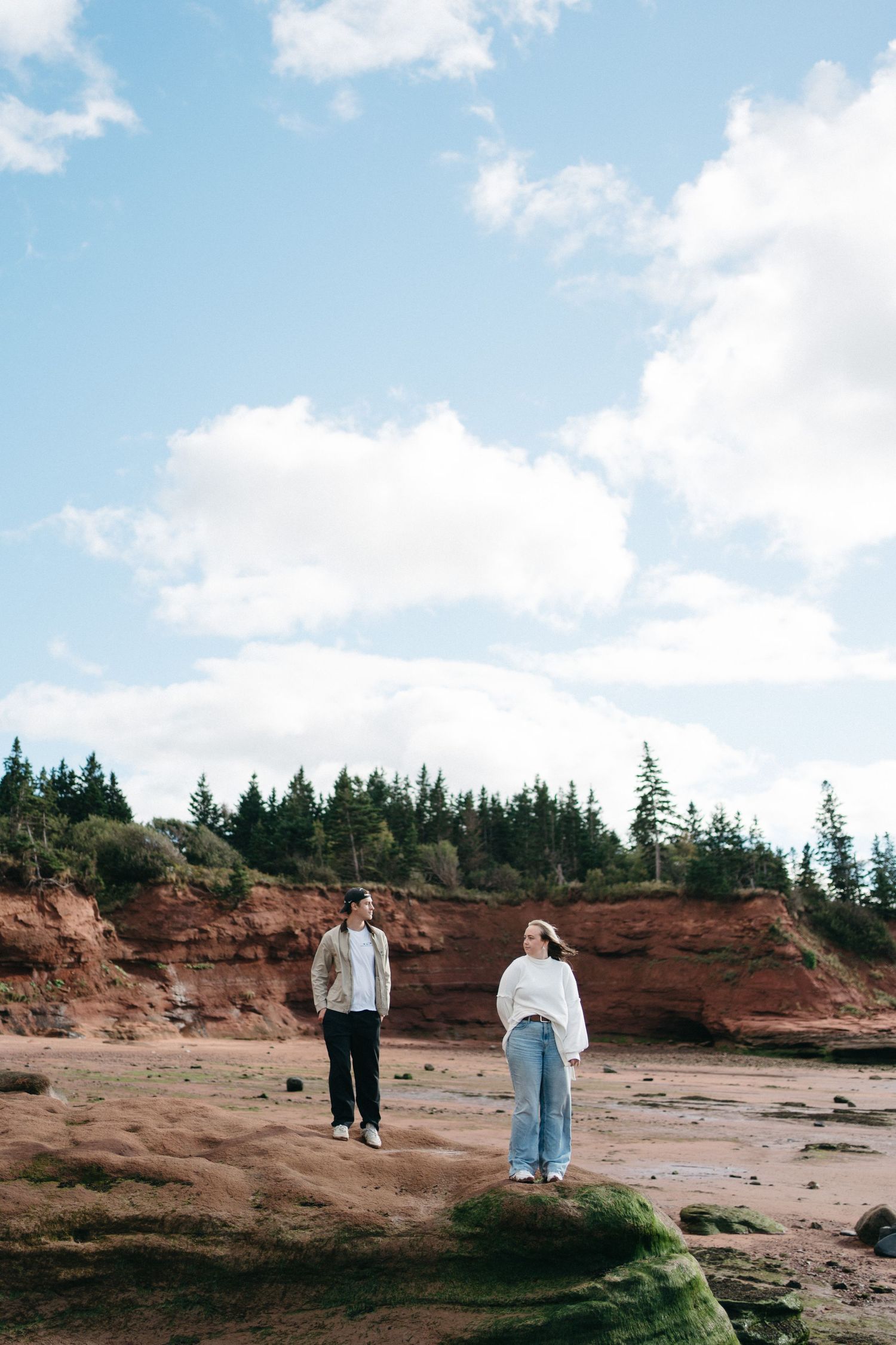 Two people stroll along a scenic coastal beach with red cliffs and evergreen trees in the background in Nova Scotia.