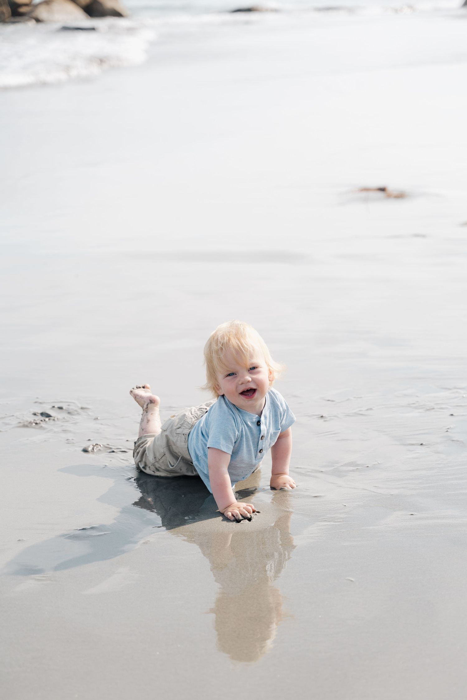 Small child wearing light blue shirt crawls and plays on wet sand at the beach, creating reflections in shallow water in Halifax, NS.