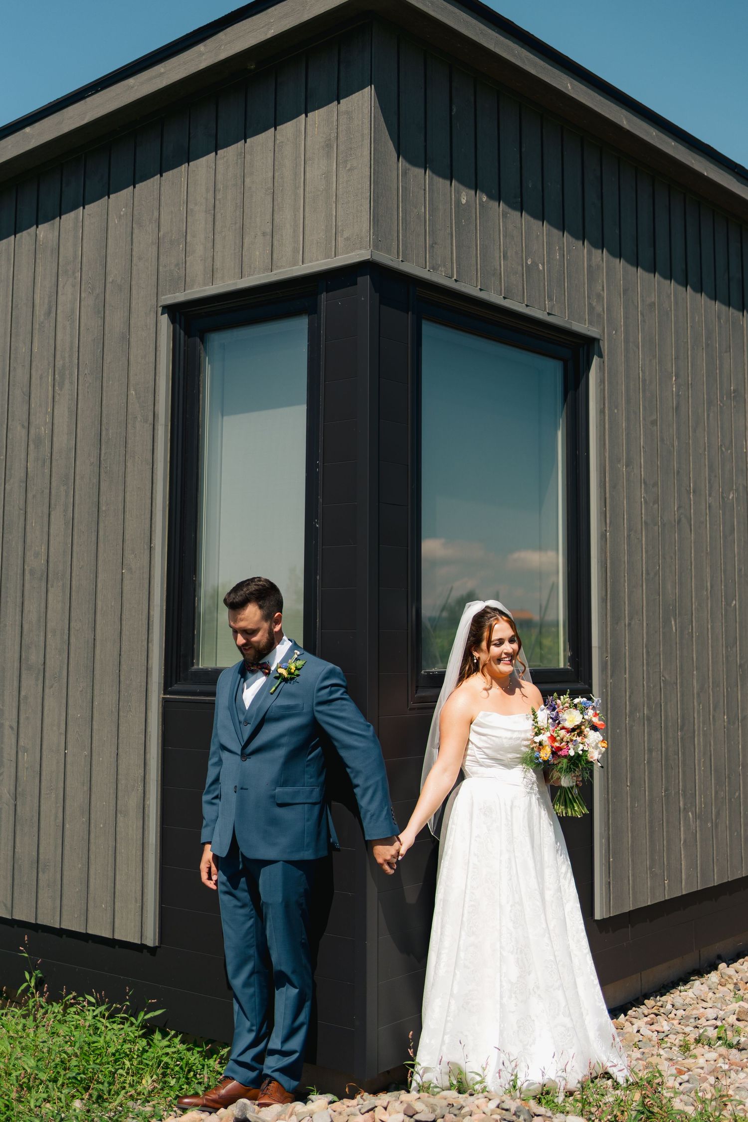 A wedding couple walks together against a modern gray barn-style building on a sunny day in Halifax, Nova Scotia.