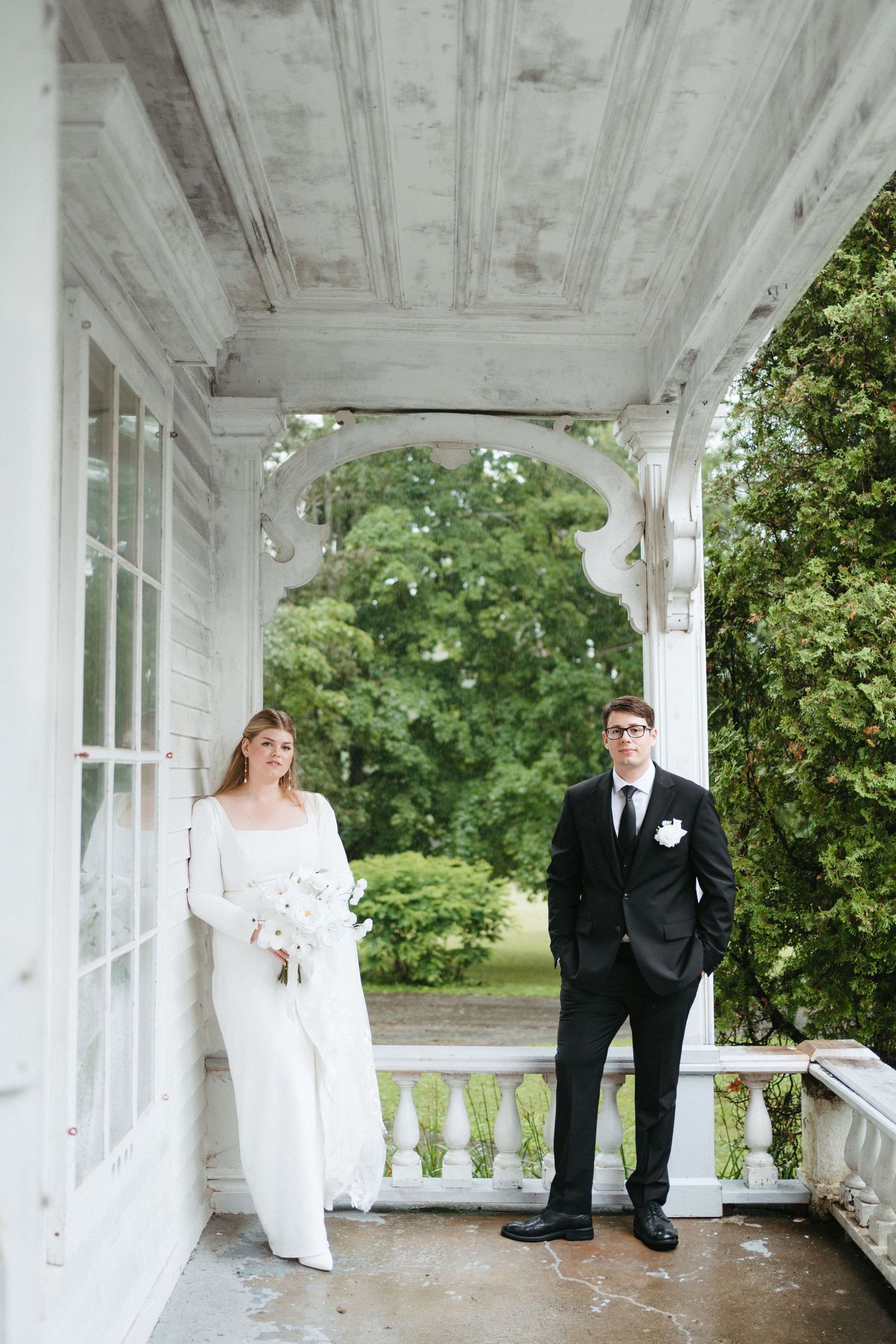A couple poses on a white columned porch with greenery in the background during their wedding day in Halifax, Nova Scotia.