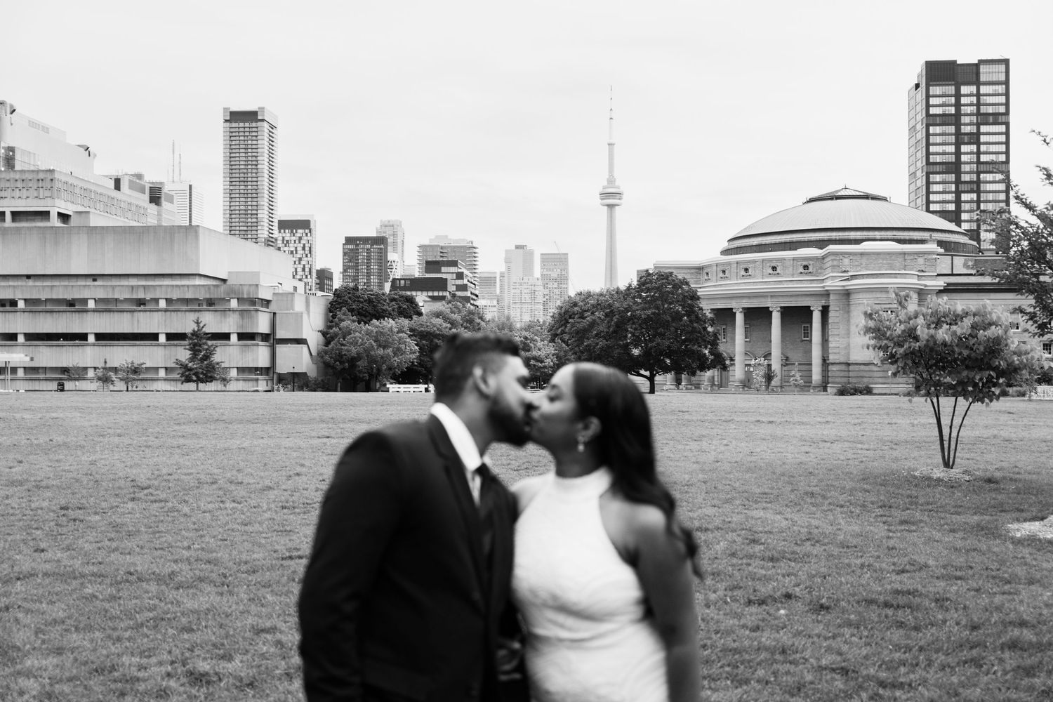 A couple shares a romantic moment on a lawn with the Toronto skyline and CN Tower visible in the background.