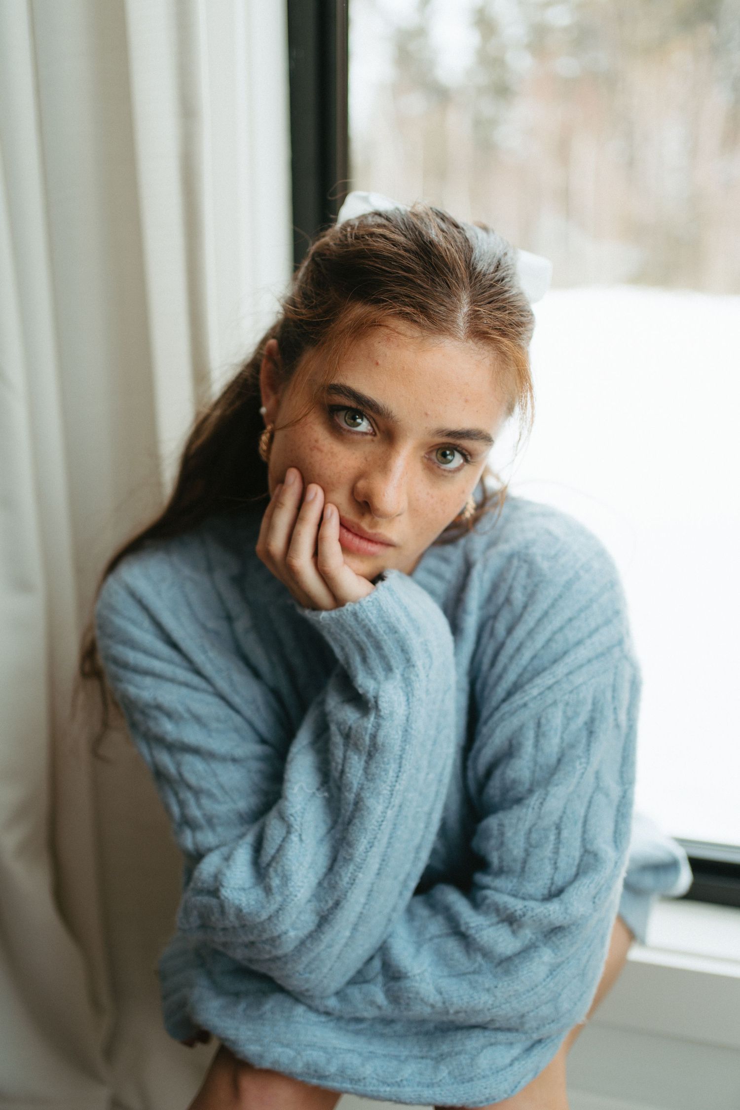Someone wearing a cozy blue sweater sits pensively by a window with natural lighting in a moody portrait in Halifax, Nova Scotia.