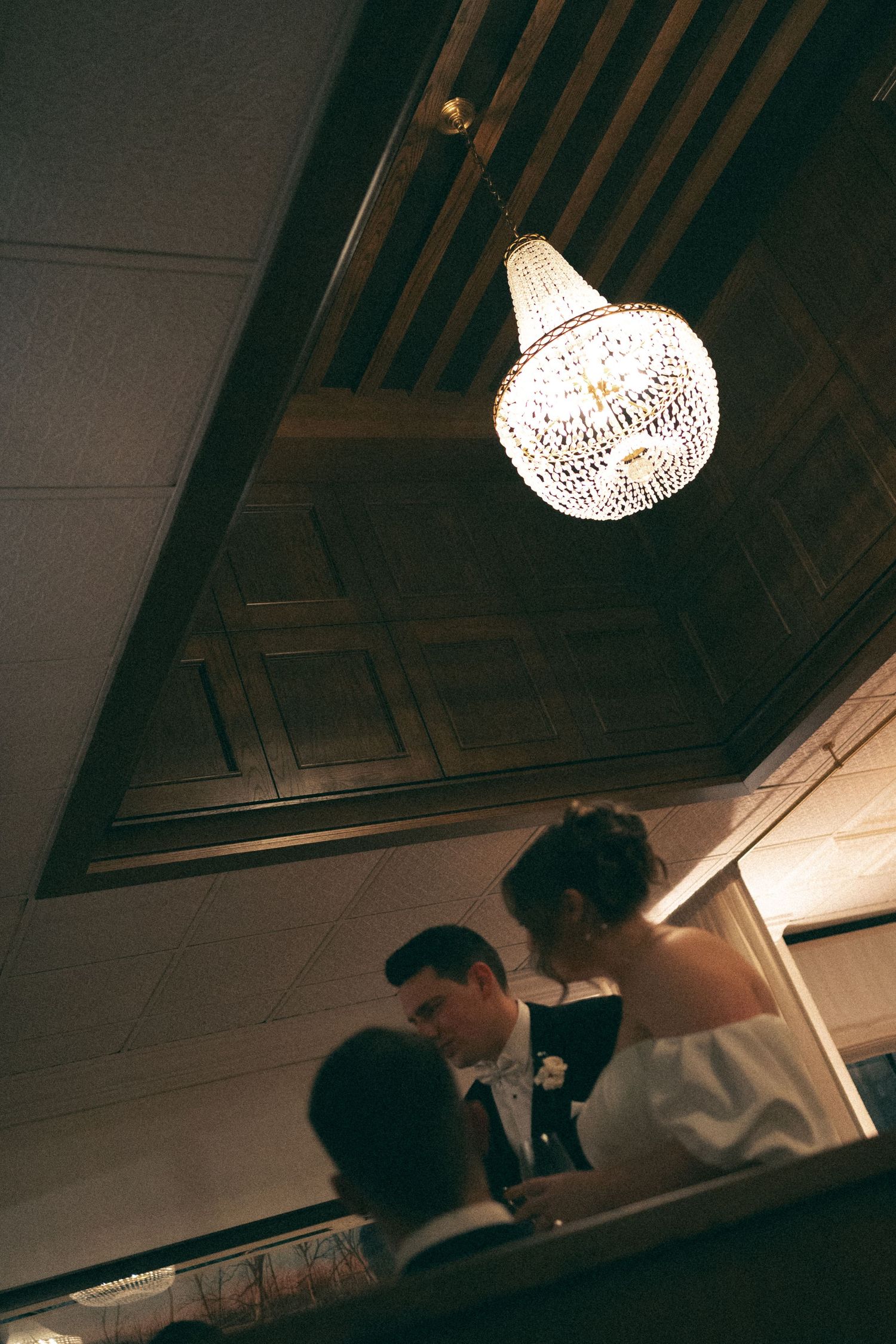 An elegant crystal chandelier hangs from a dark wooden ceiling above wedding guests at Lord Nelson Hotel, Halifax, Nova Scotia.