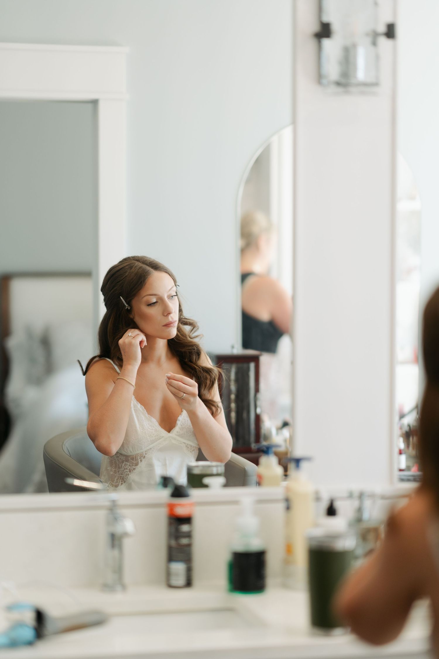 A bride gets ready in front of a bright bathroom mirror with makeup products visible on the counter in Halifax, Nova Scotia.