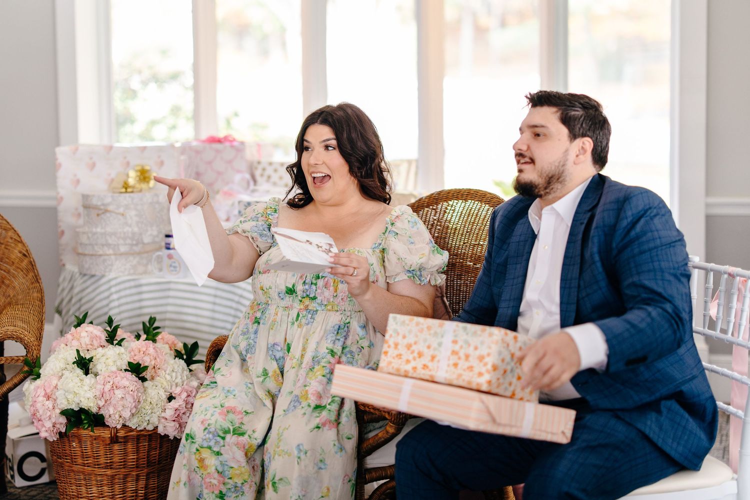 Couple opening gifts at bridal shower, woman in floral dress looking surprised.