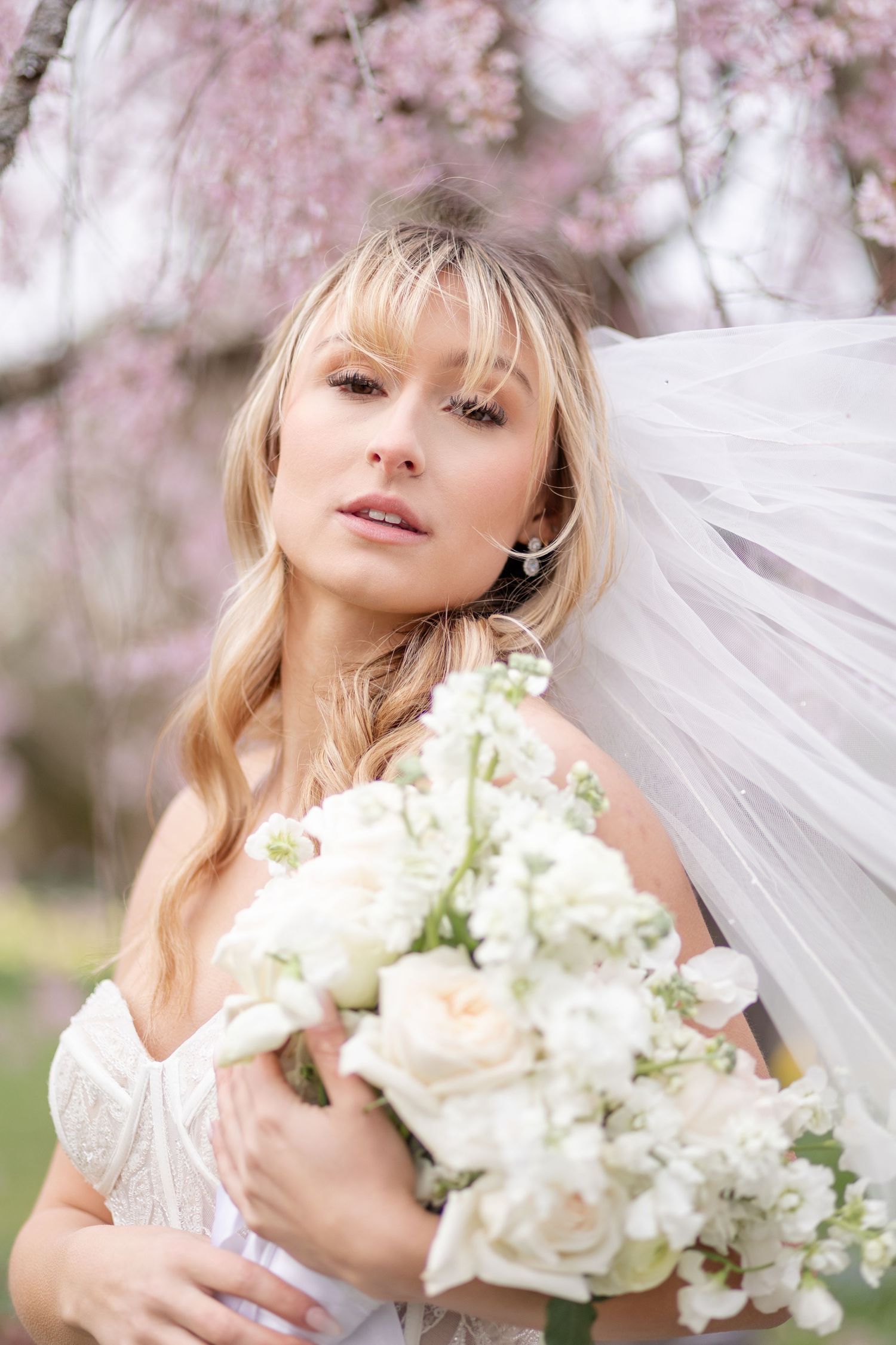 Blonde bride with veil holds white bouquet under pink cherry blossoms in spring.