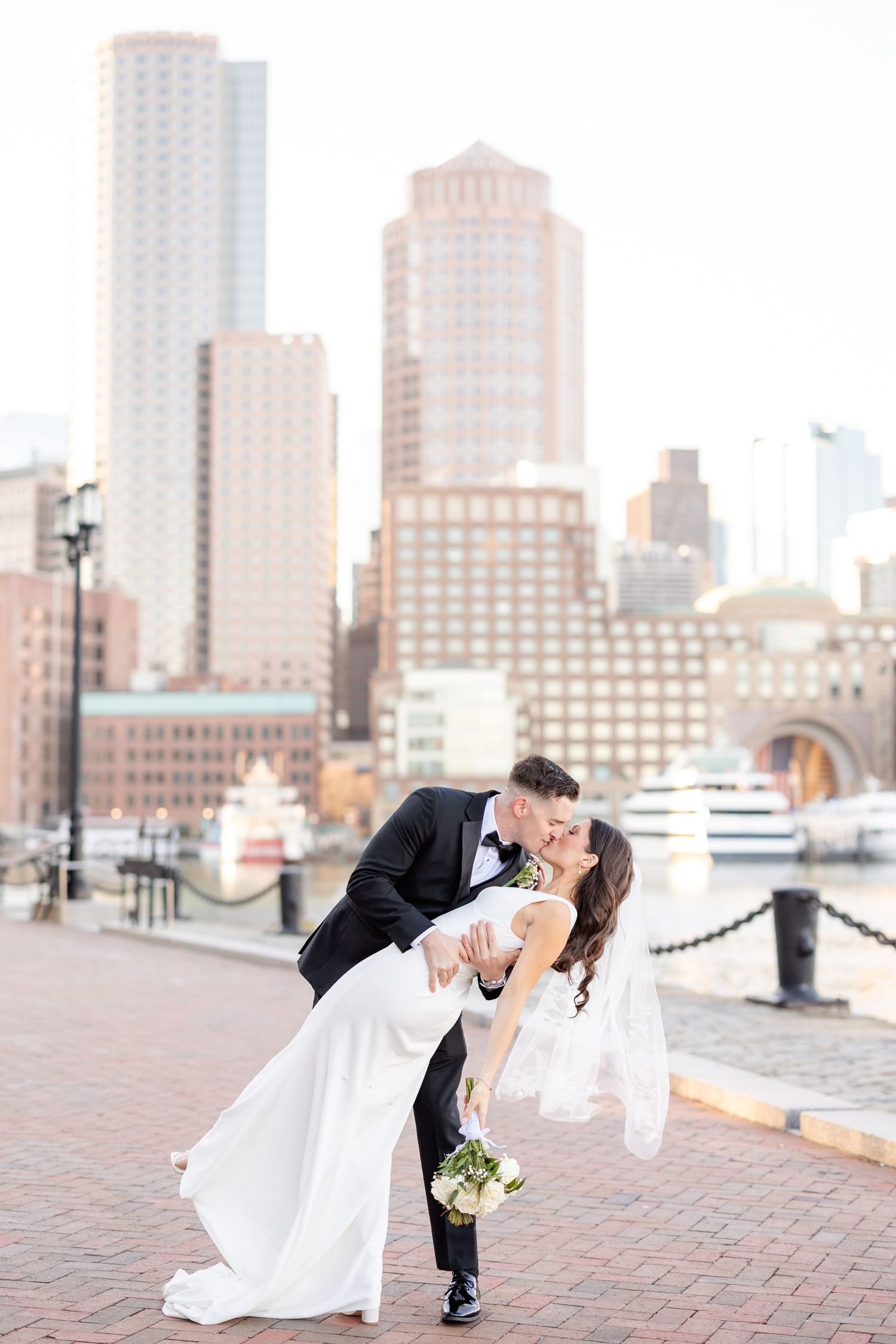 Groom dips bride for a kiss with Boston city skyline and harbor beautifully framed behind them.