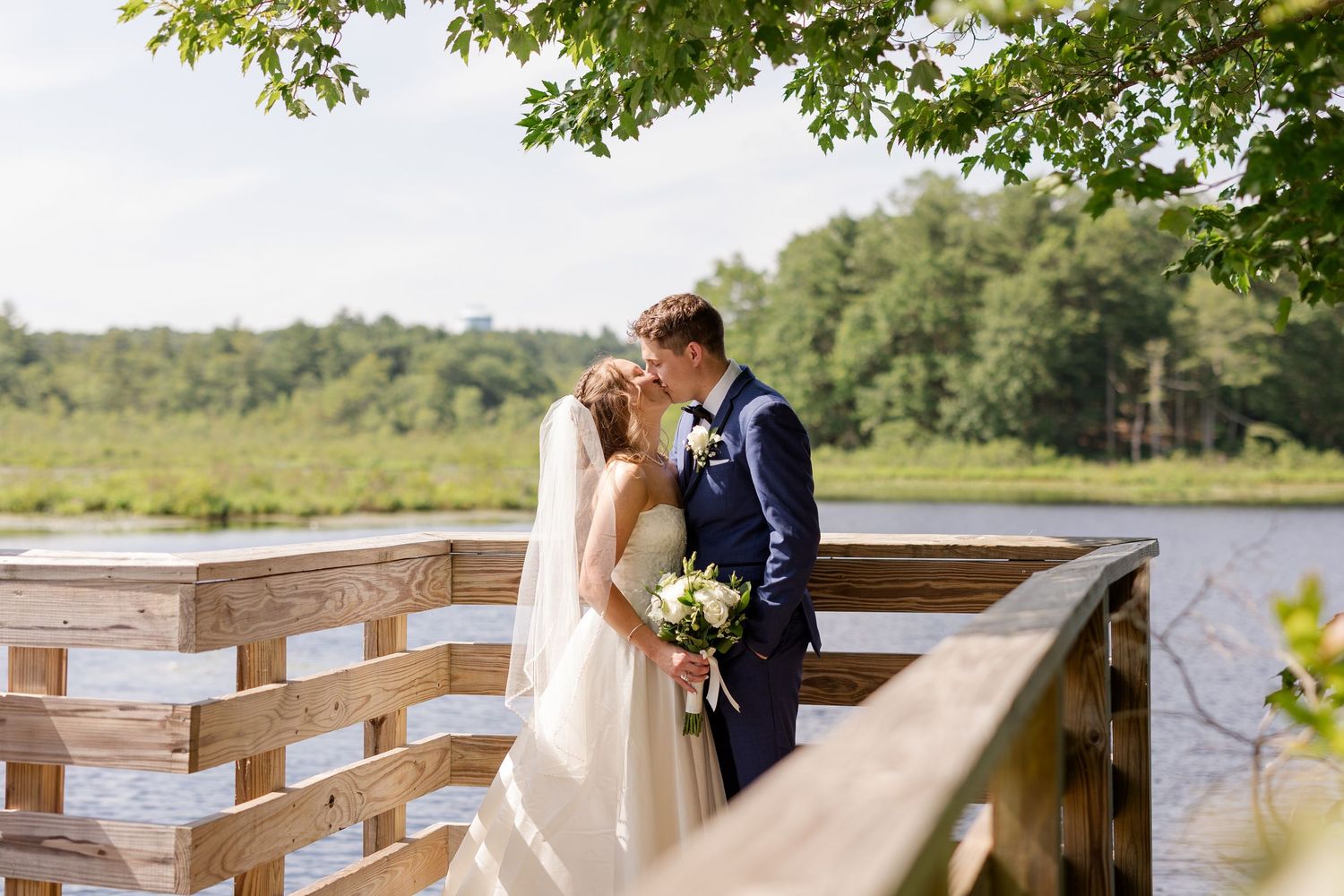 Wedding couple shares a tender kiss on a wooden deck overlooking a serene lake and green forest backdrop.