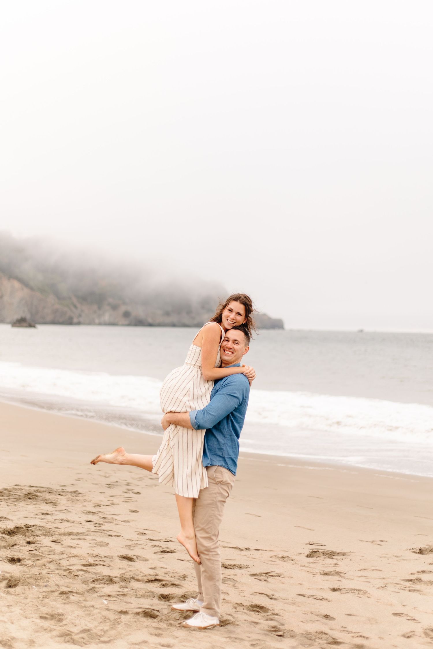 Happy couple sharing a joyful hug on a foggy beach, woman lifted off the ground smiling.