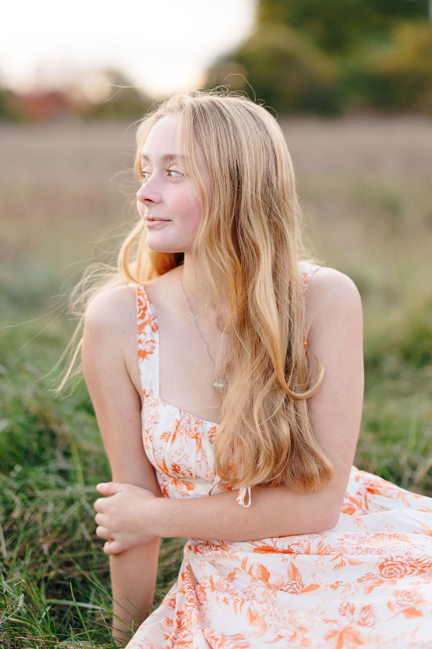 Blonde teen girl in orange floral sundress sitting in grassy field at golden hour.