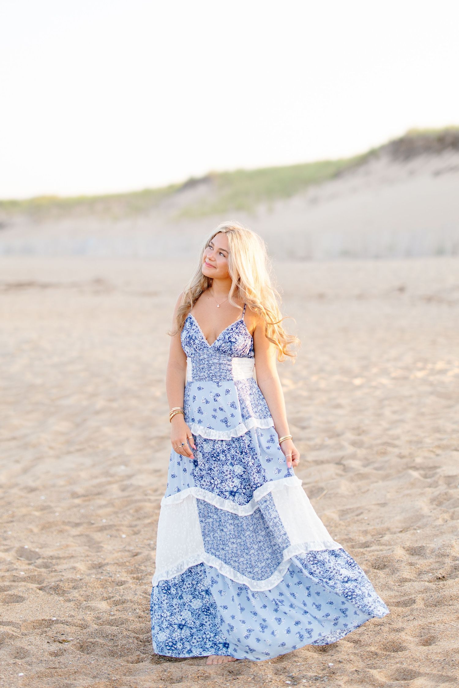 Blonde teenage girl in blue floral maxi dress posing on a sandy beach at sunset.