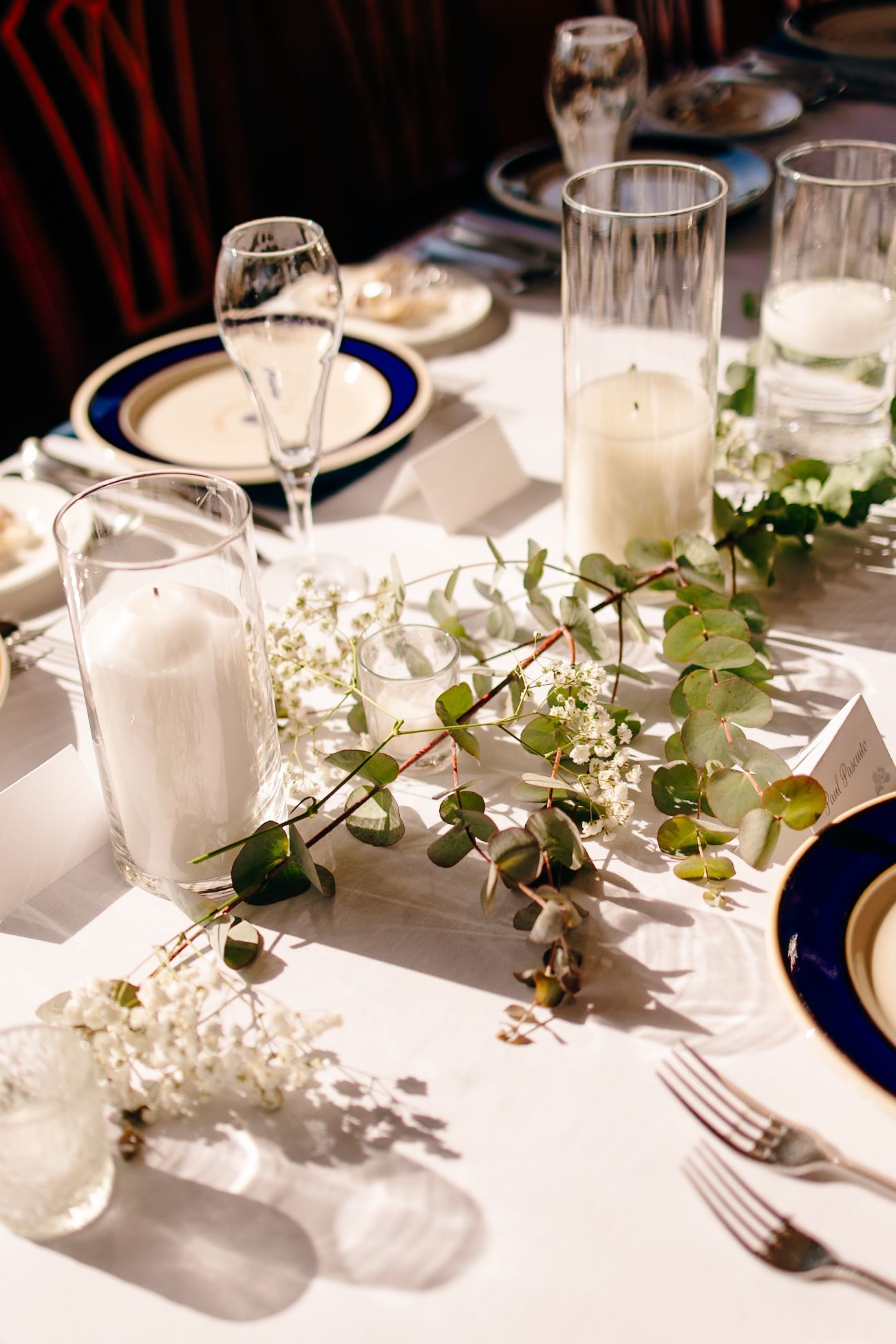 Elegant wedding table setting with white candles, eucalyptus greenery, blue-rimmed plates, and crystal glassware on white linen.