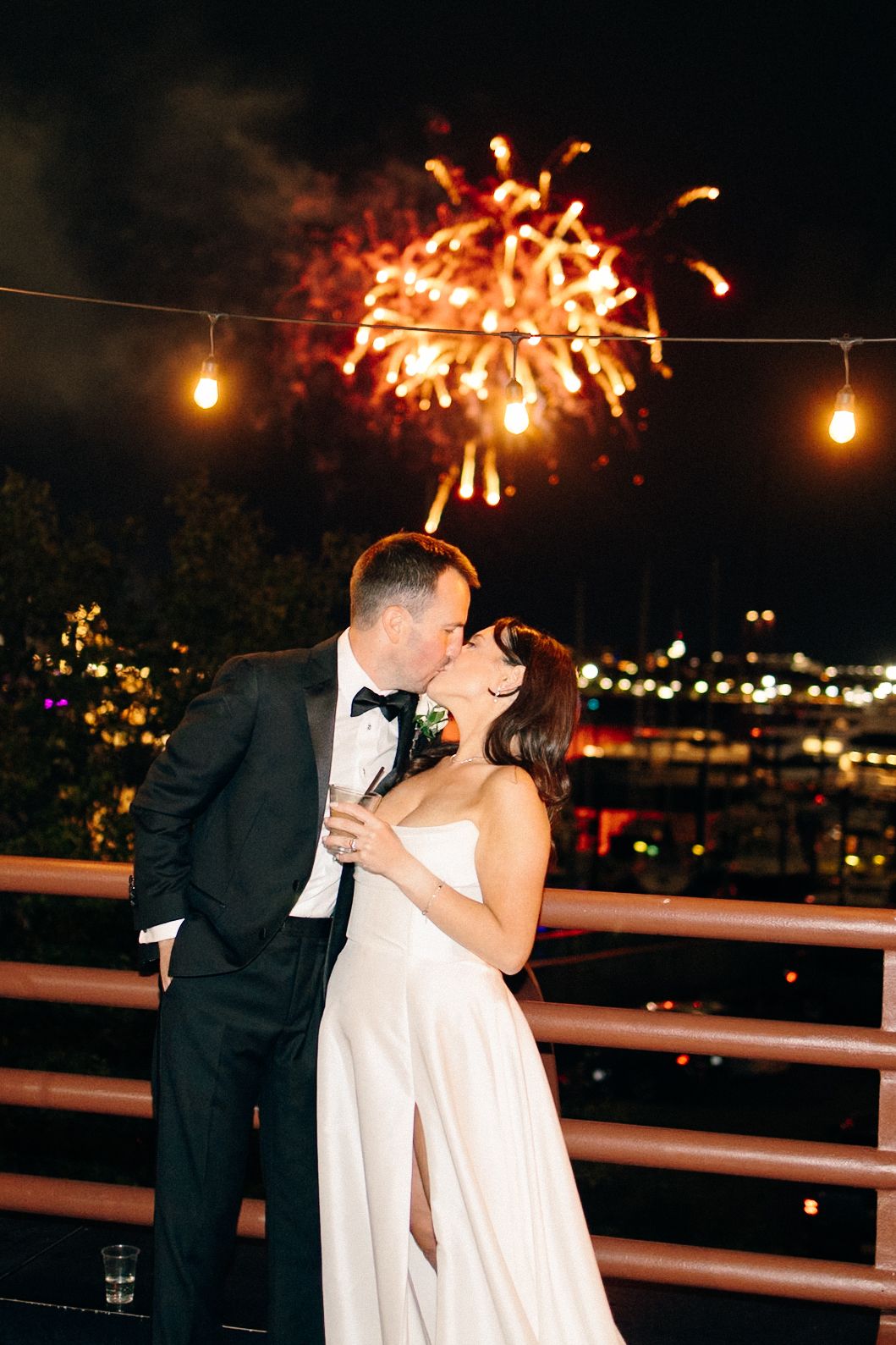 Bride and groom share a kiss on outdoor terrace with colorful fireworks bursting in night sky.