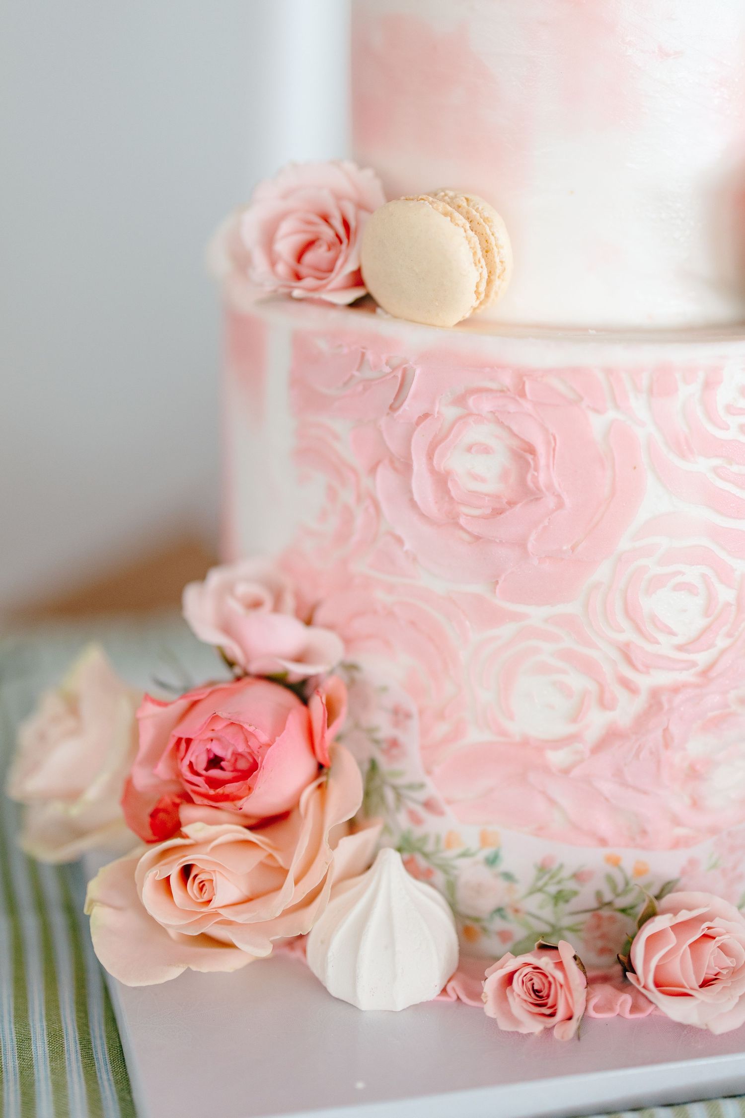 Elegant pink wedding cake decorated with rose frosting, fresh roses, meringues, and a macaron on top.
