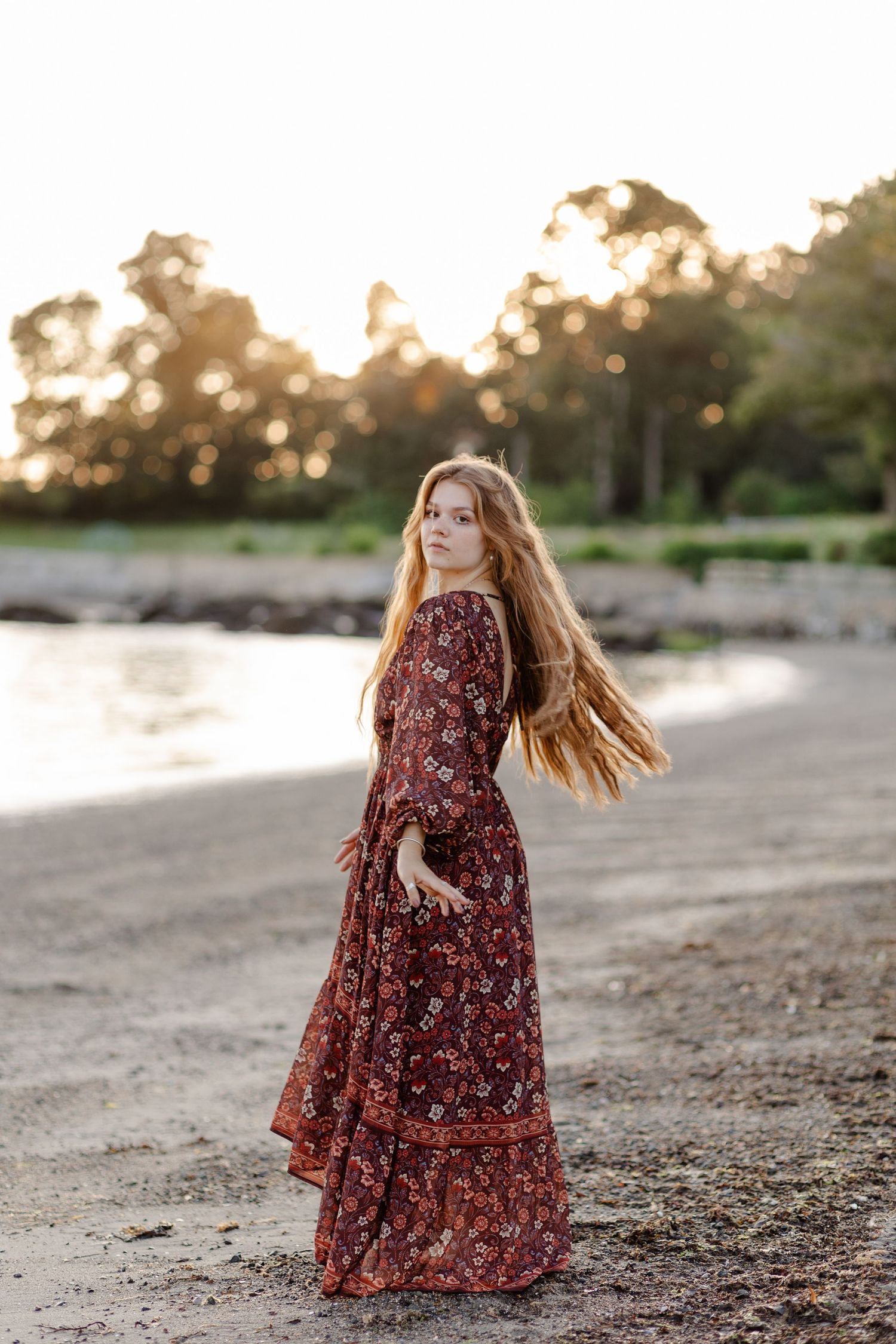 Redhead woman in long burgundy floral dress posing on a beach shore at sunset.