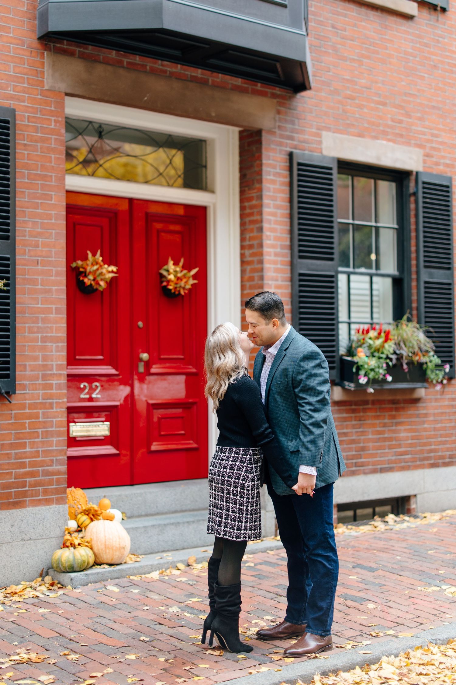 Couple sharing a kiss in front of red door at number 22, surrounded by fall pumpkins and leaves.