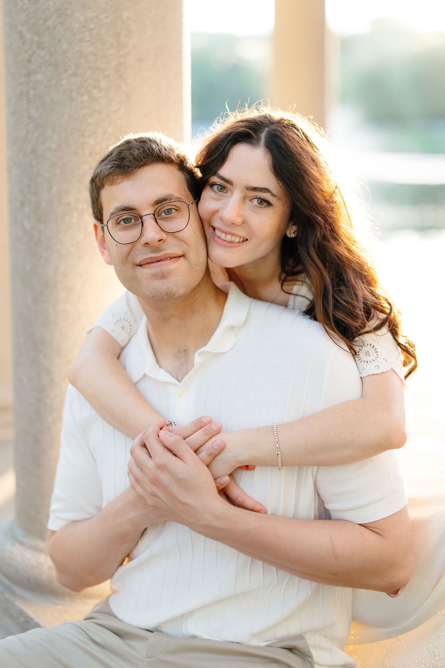 A smiling couple embraces outdoors, woman with dark wavy hair hugging man in glasses near stone columns.