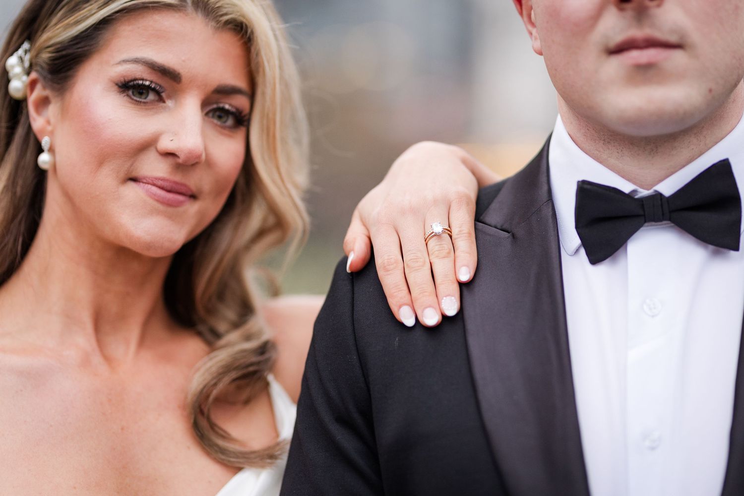 Bride showing diamond engagement ring while standing beside groom in black tuxedo.