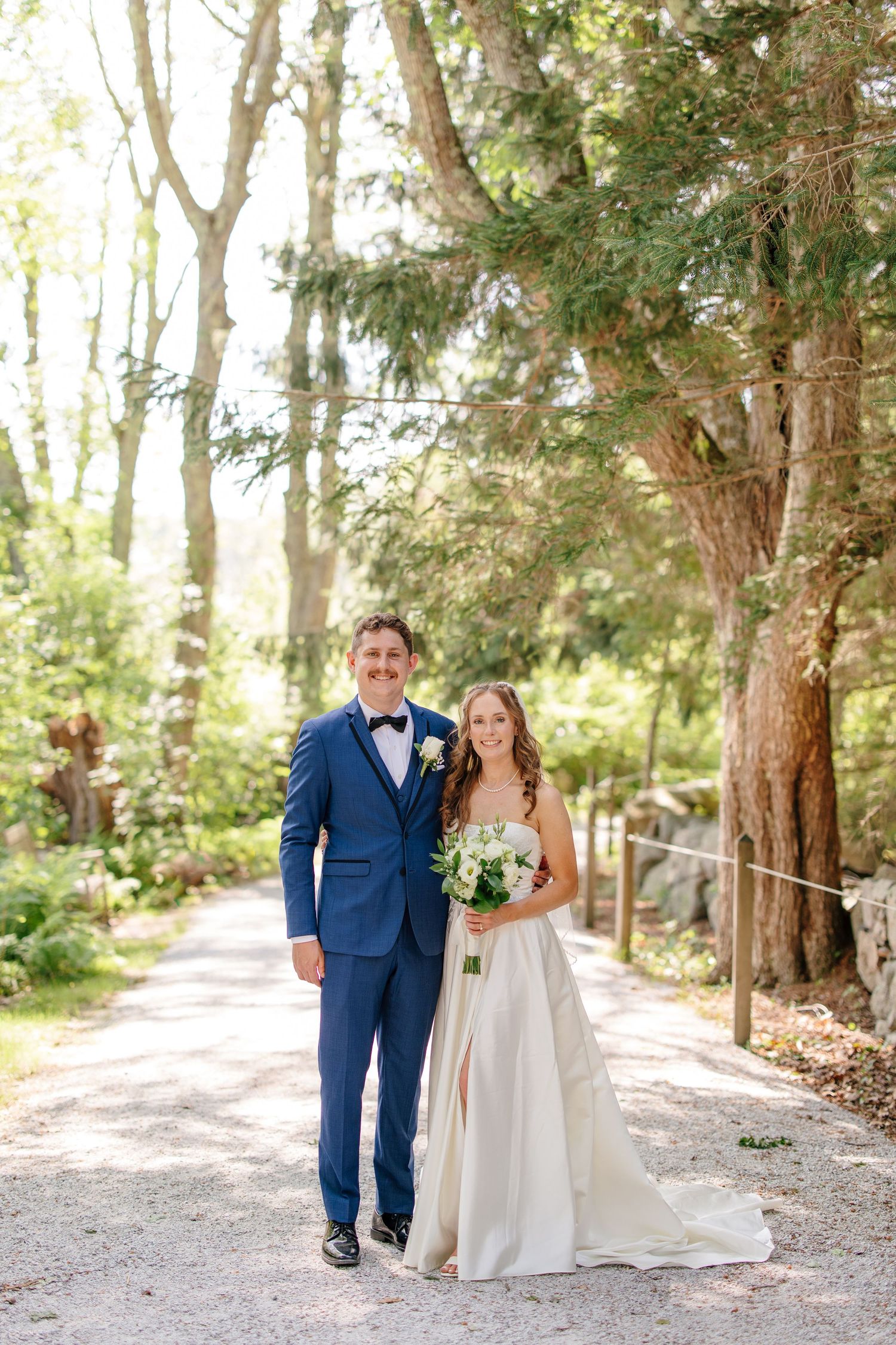 Bride and groom pose together on a sunlit forest path, bride holding a white bouquet in elegant gown.