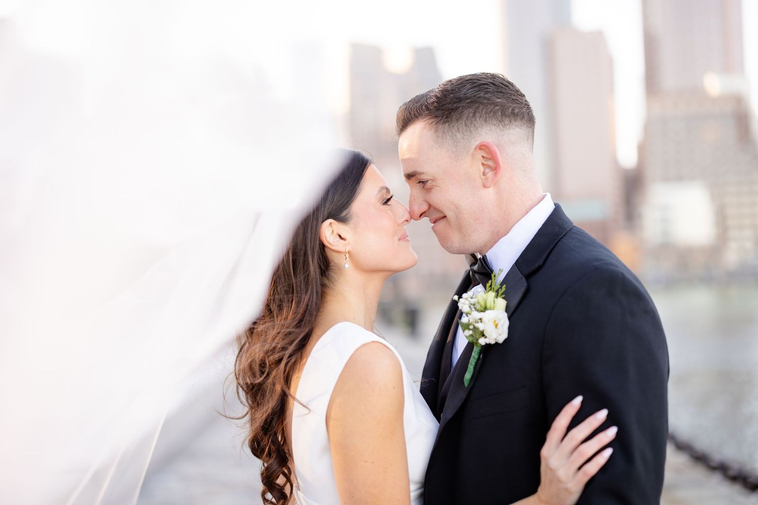 Bride and groom share an intimate almost-kiss moment with veil flowing, city skyline softly blurred in background.
