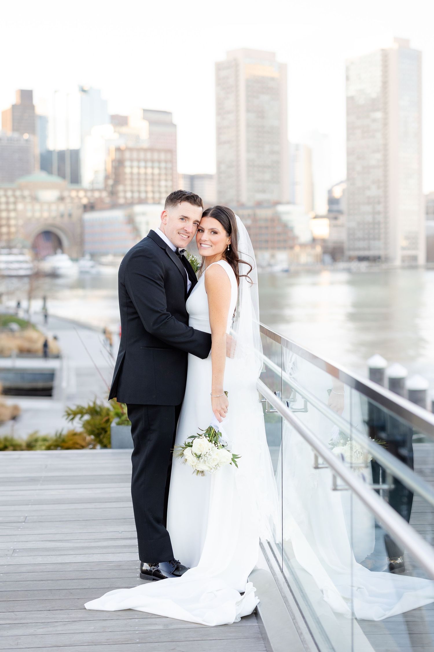 Bride and groom embrace smiling on waterfront deck with Boston harbor and city skyline behind them.