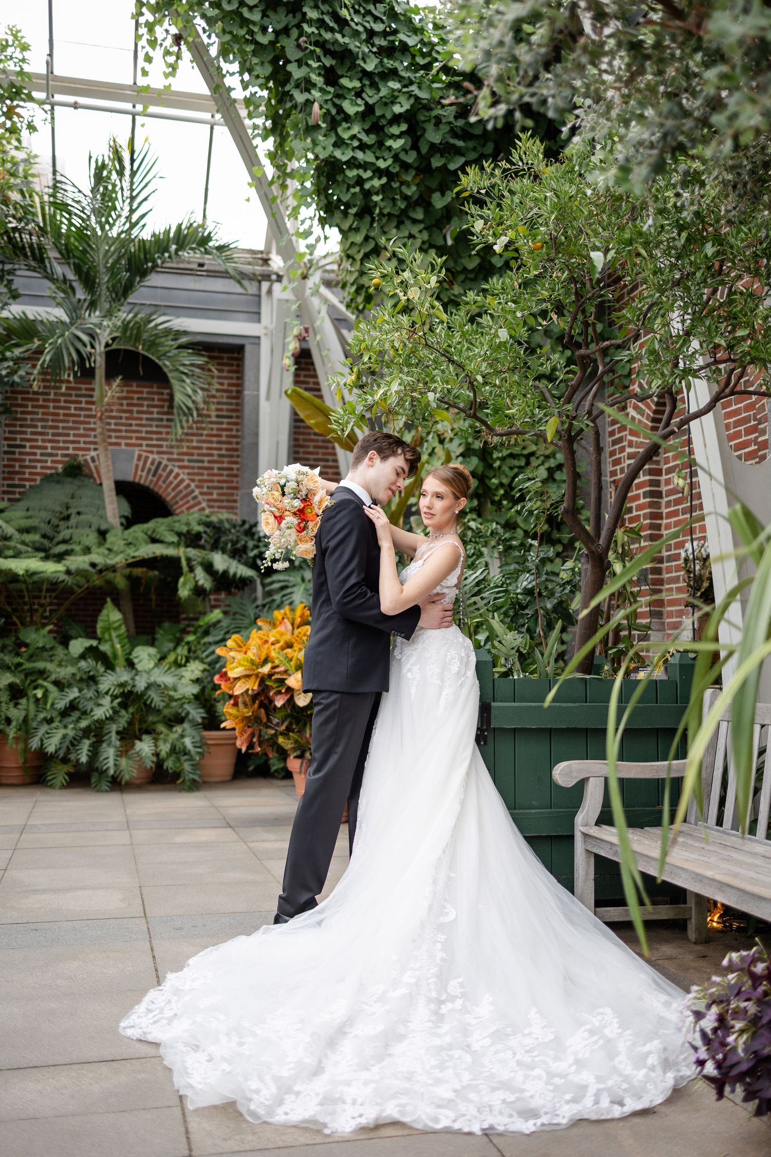 Couple embraces in lush garden courtyard, bride in lace gown holding colorful bouquet.