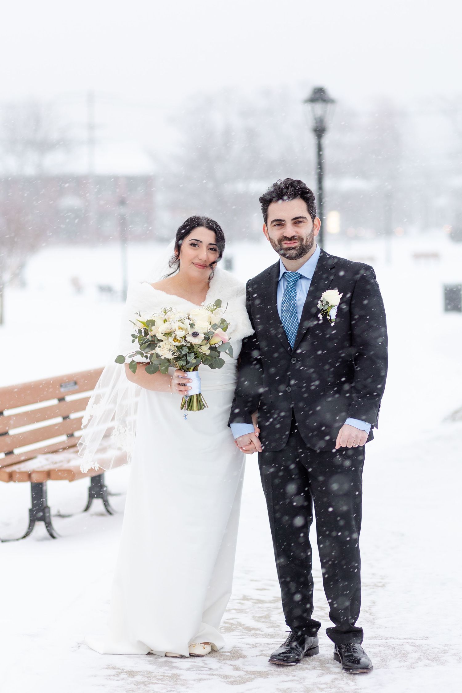 Bride and groom holding hands in snowy winter park, bride holding white bouquet in elegant white gown.