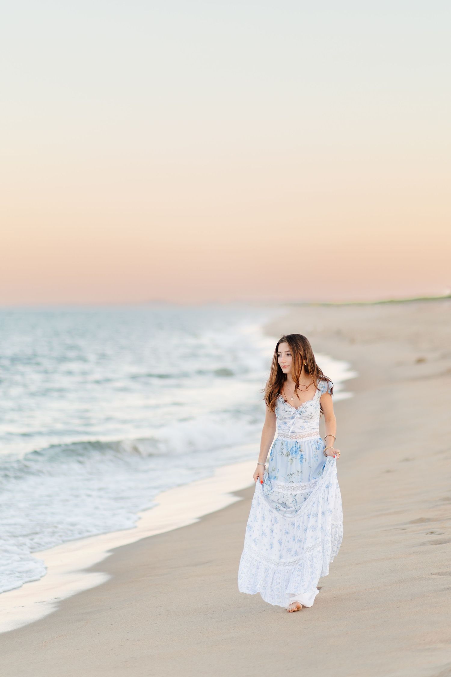 Young woman in white floral maxi dress walking on beach at sunset.